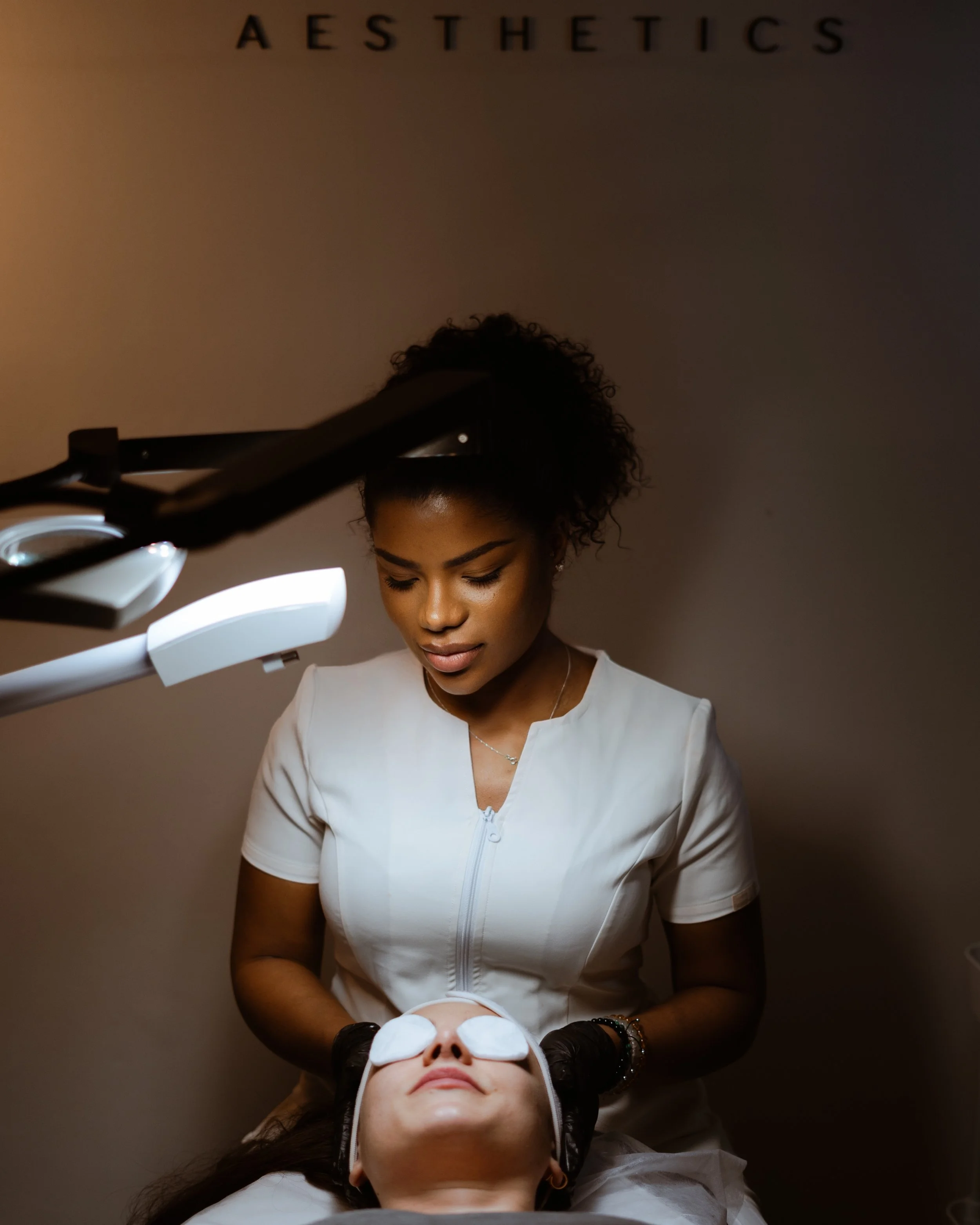 Aesthetic skincare treatment. A woman in a white uniform performs a facial on a client lying down with eye pads. The background has the word 'AESTHETICS' on the wall.