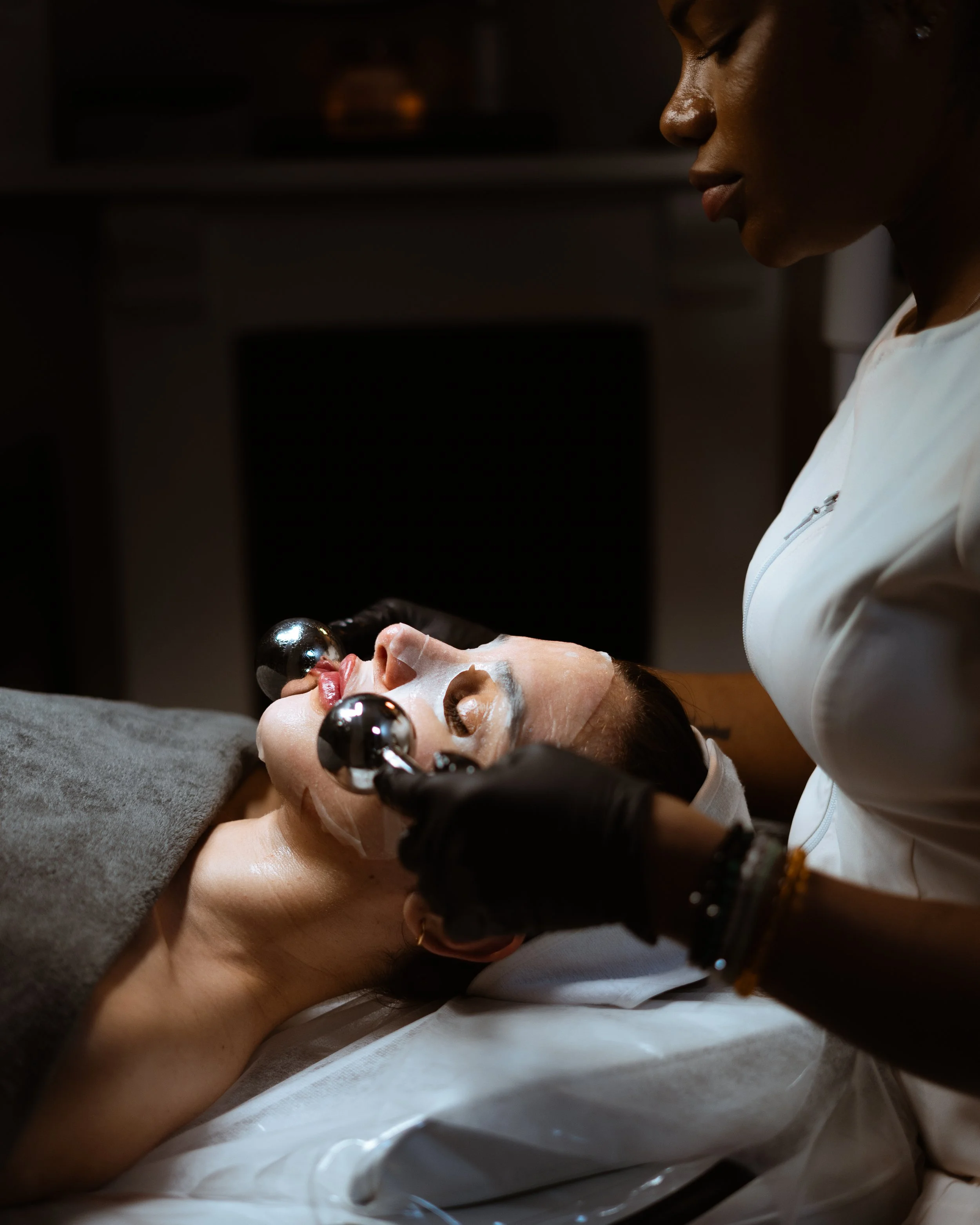 A woman is lying on a spa bed receiving a facial treatment from an esthetician, who is applying a facial mask and using massage tools.