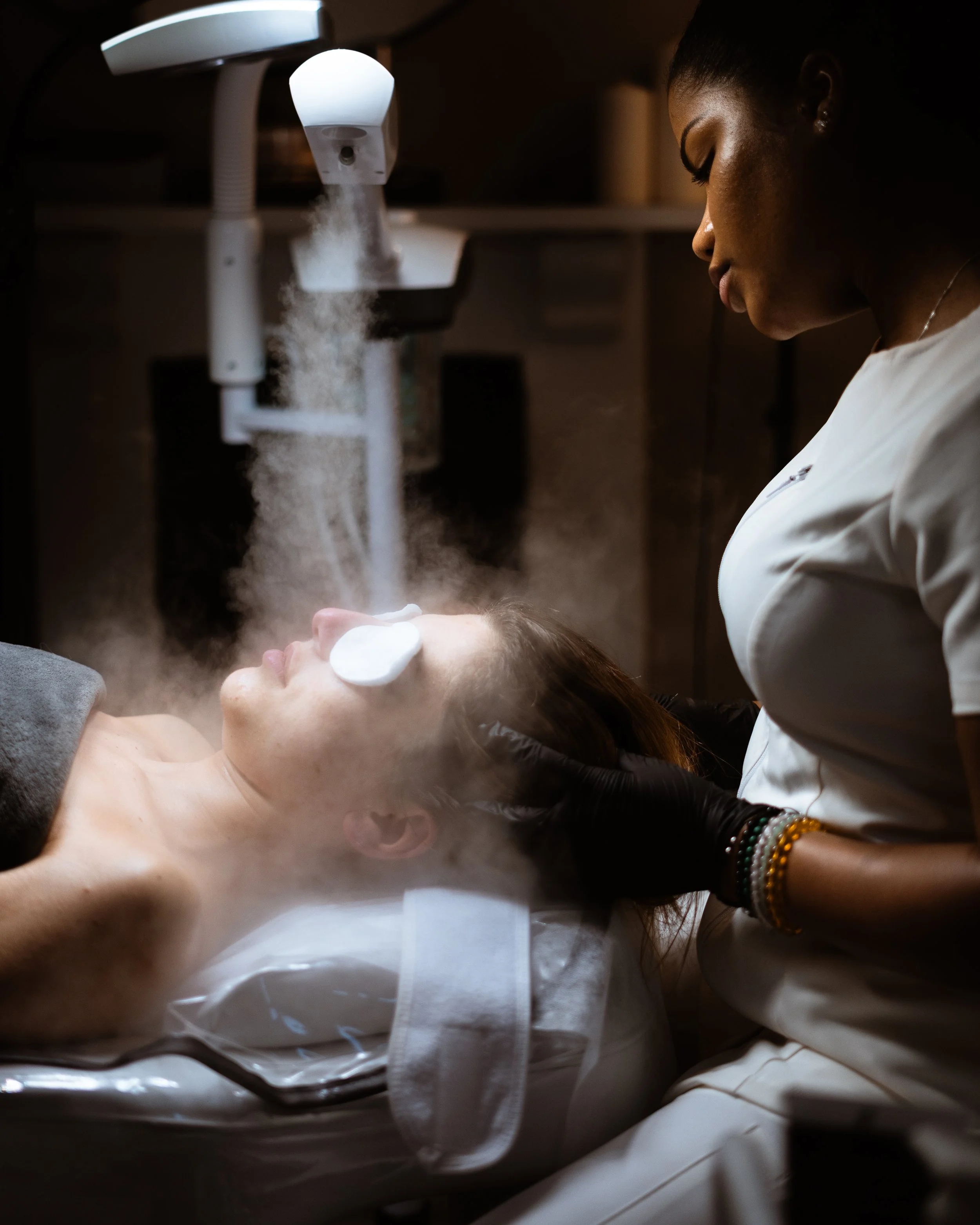 A woman is lying on a treatment bed with protective eye covers on her eyes, receiving a facial treatment from a skincare professional in a clinical setting with steam and medical equipment.