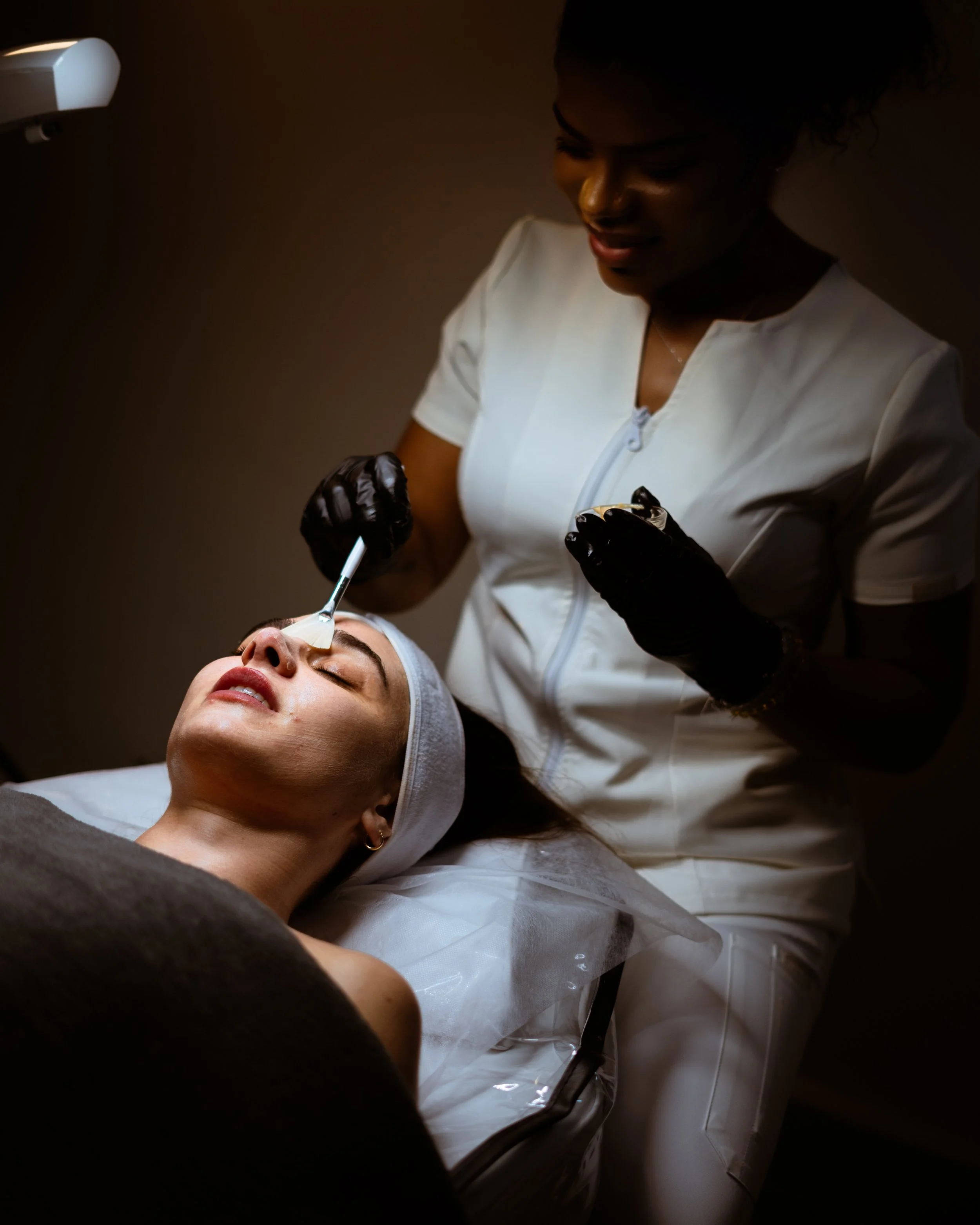 A woman lying on a treatment bed receiving a facial treatment from a skincare professional using a brush on her face.