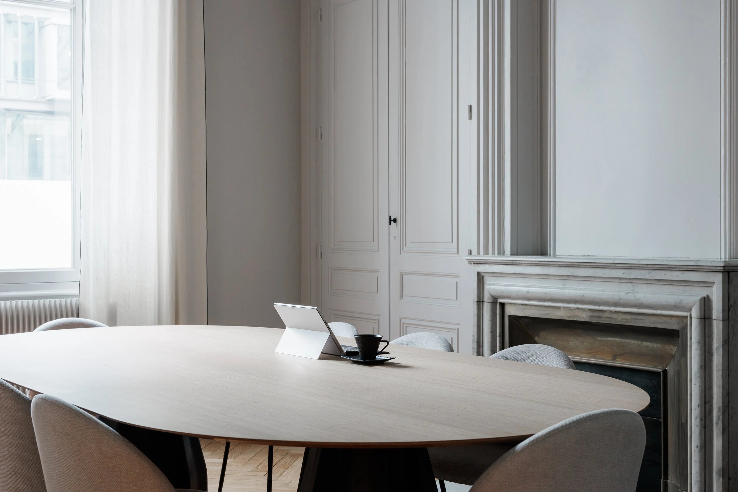 Salle de réunion avec table ovale, six chaises, un ordinateur portable, une tasse, dans une pièce lumineuse avec cheminée en marbre et grandes fenêtres