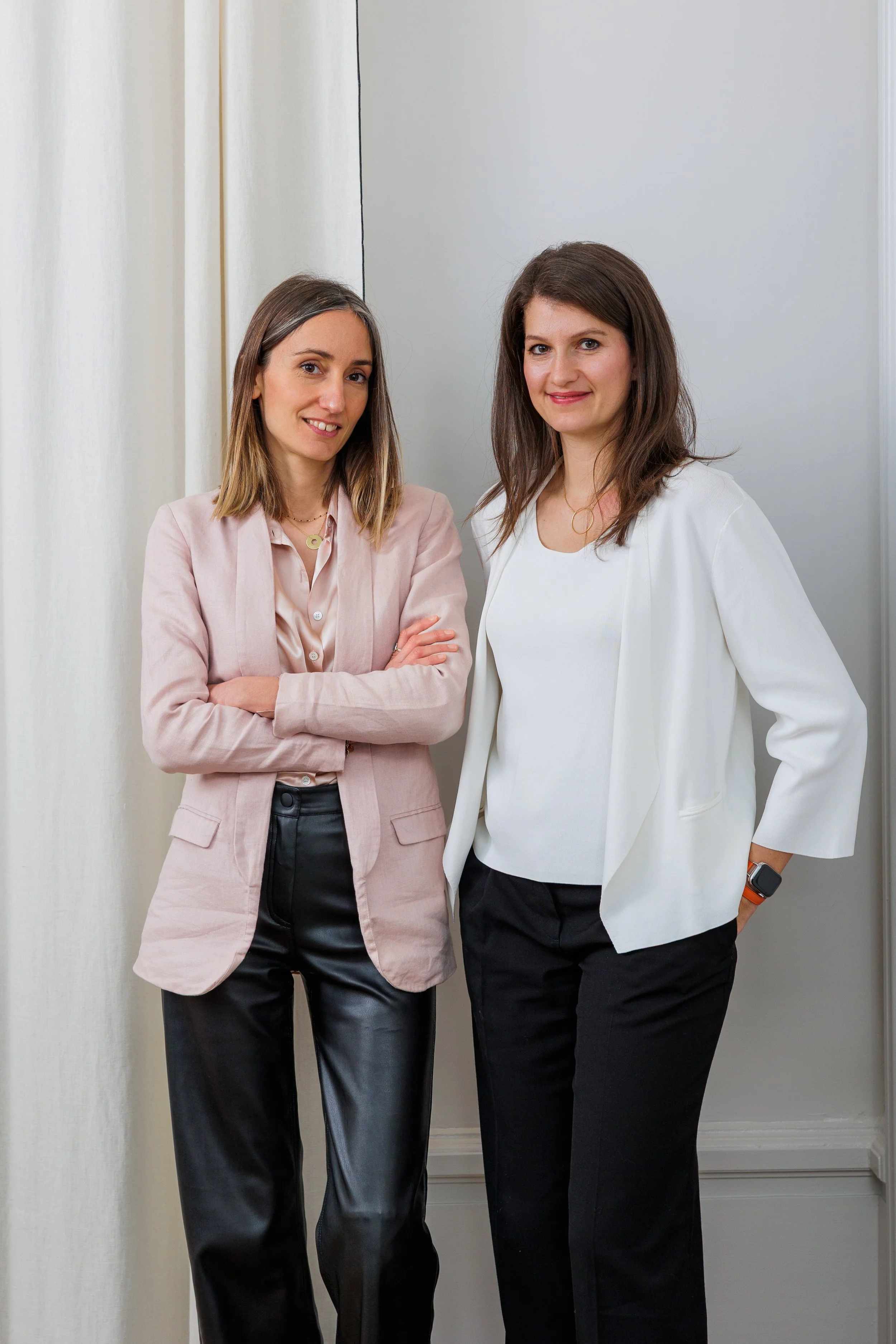 Deux femmes posent côte à côte dans une pièce, l'une avec un blazer rose et pantalon en simili cuir, l'autre avec une veste blanche et pantalon noir