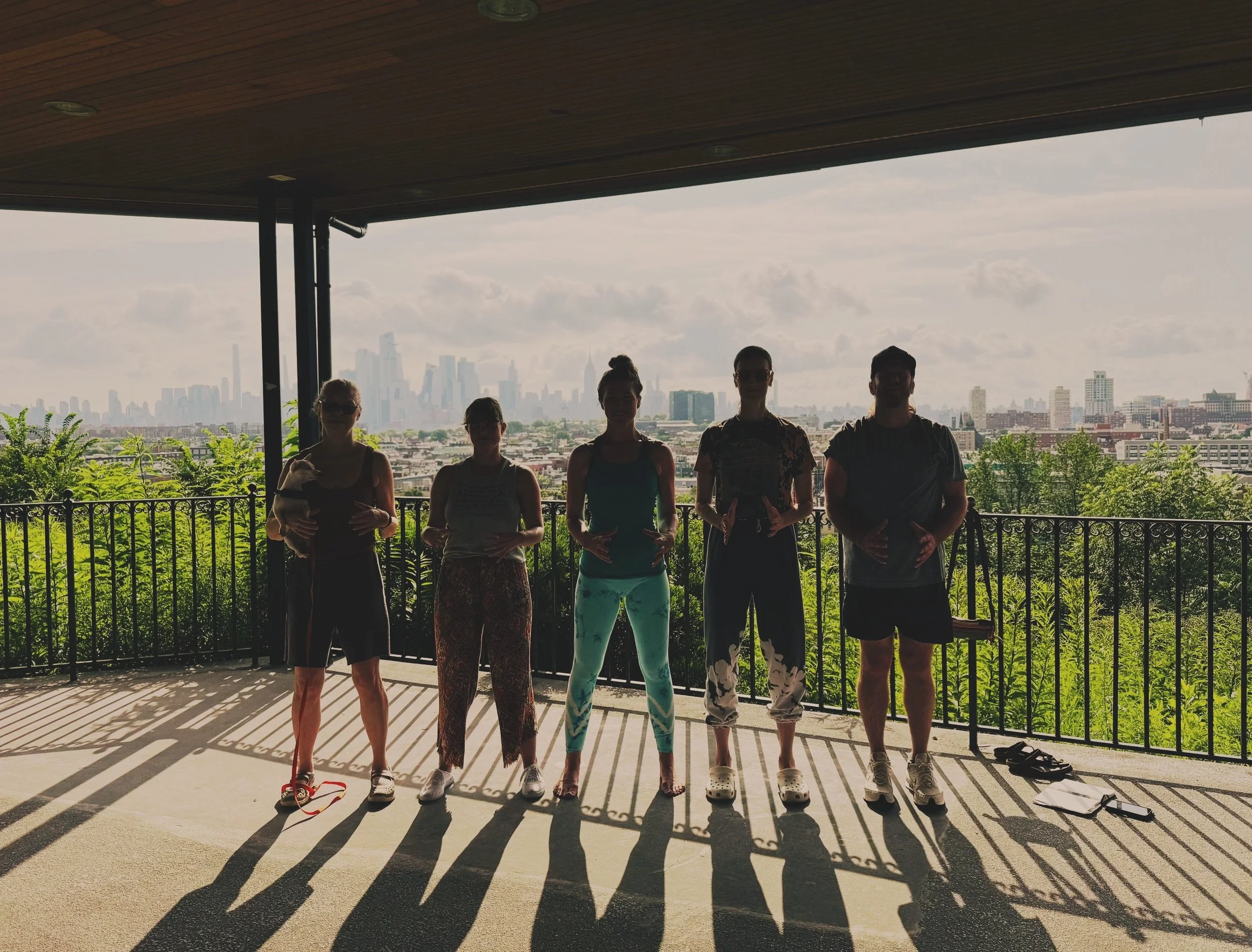 Five people stand side by side on a balcony practicing QiGong with a city skyline in the background, shadows cast on the ground.