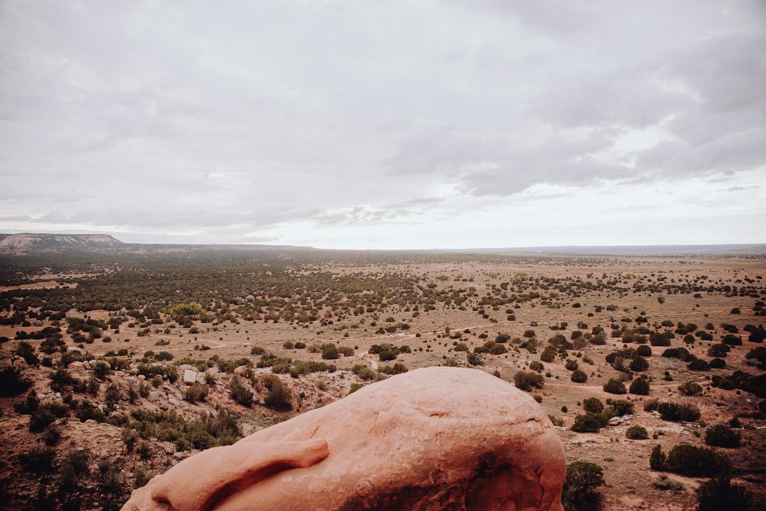 Taos New Mexico Retreat with EKAM. Wide view of a desert landscape with scattered bushes, a cloudy sky, and distant flat-topped mesas, seen from an elevated rock vantage point.