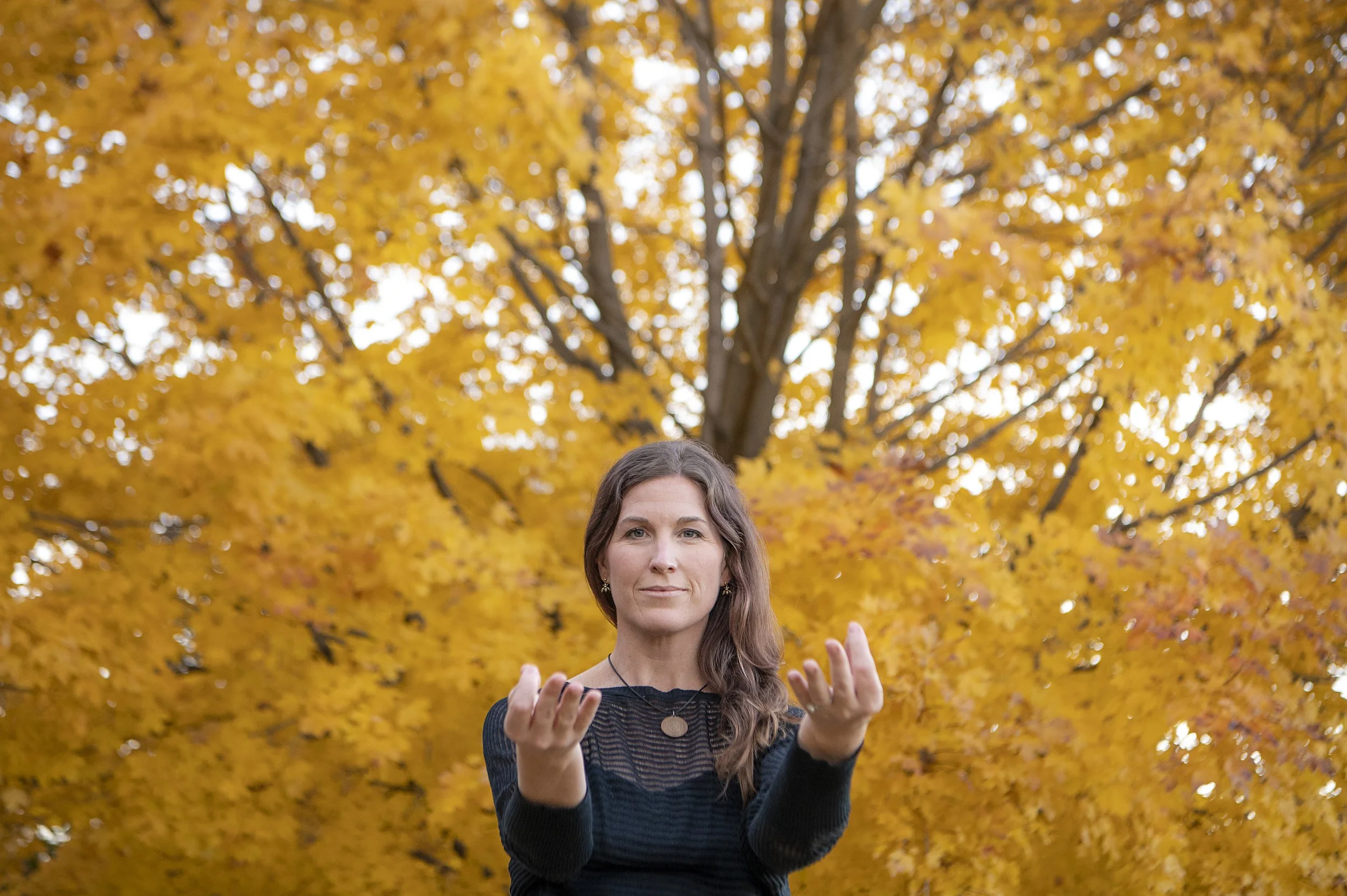 A woman standing outdoors in front of a tree with bright orange autumn leaves, with her hands raised in front of her chest practicing QiGong.
