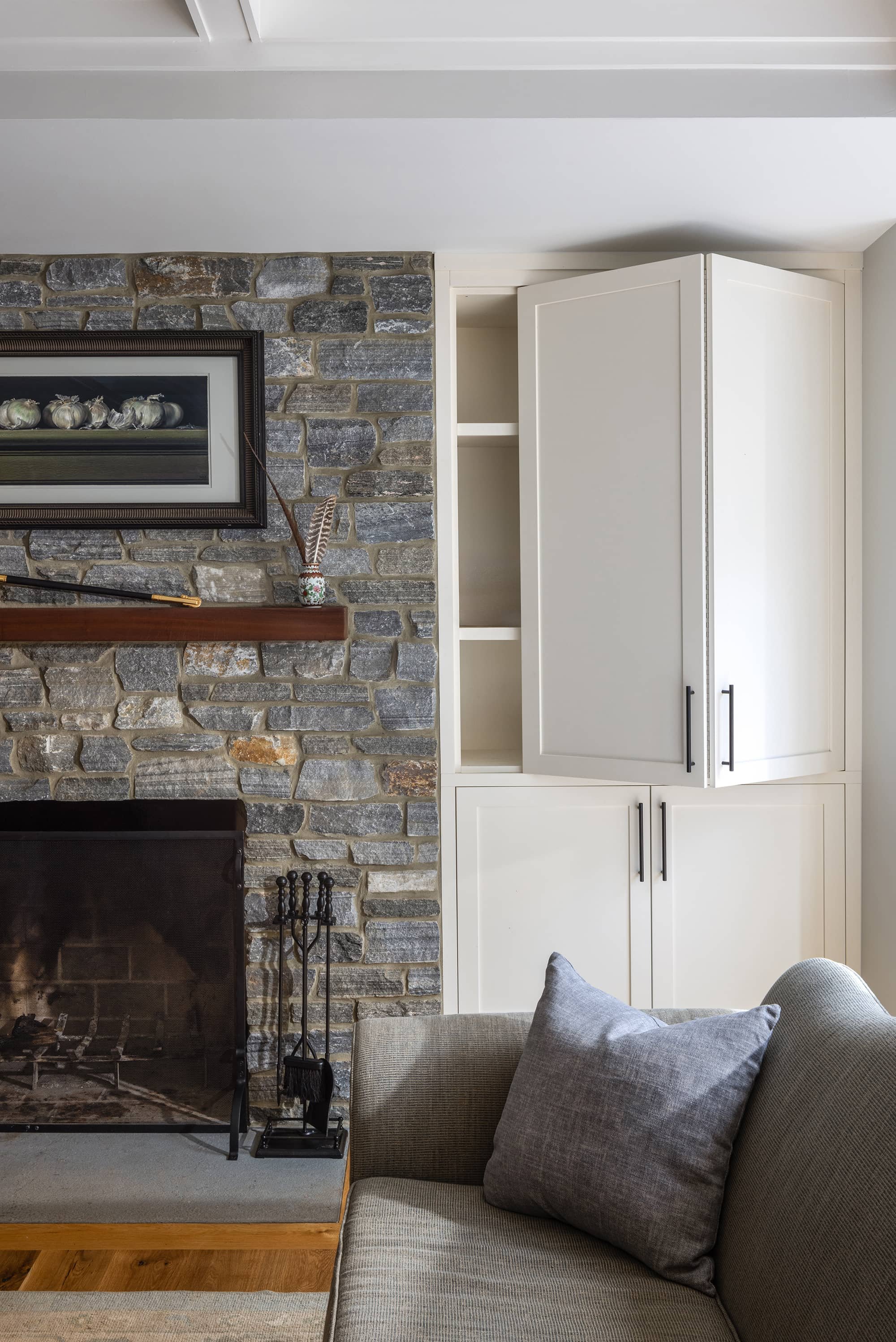 Living room with a stone fireplace, a wooden mantel with a framed painting, white built-in cabinets, and a gray sofa with a matching pillow.