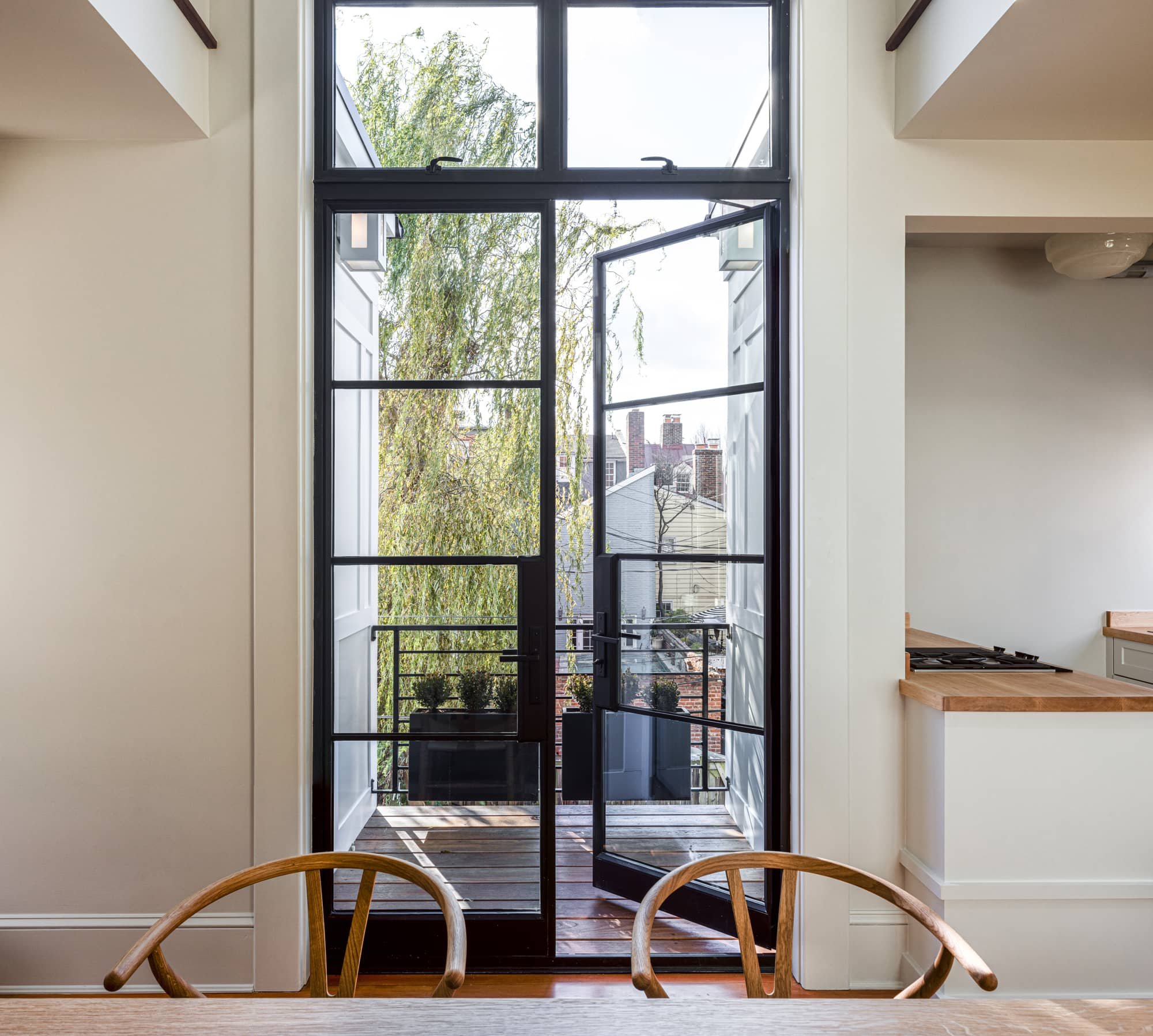 View through black-framed glass door to small balcony with potted plants and neighboring houses, with kitchen area visible to the right.