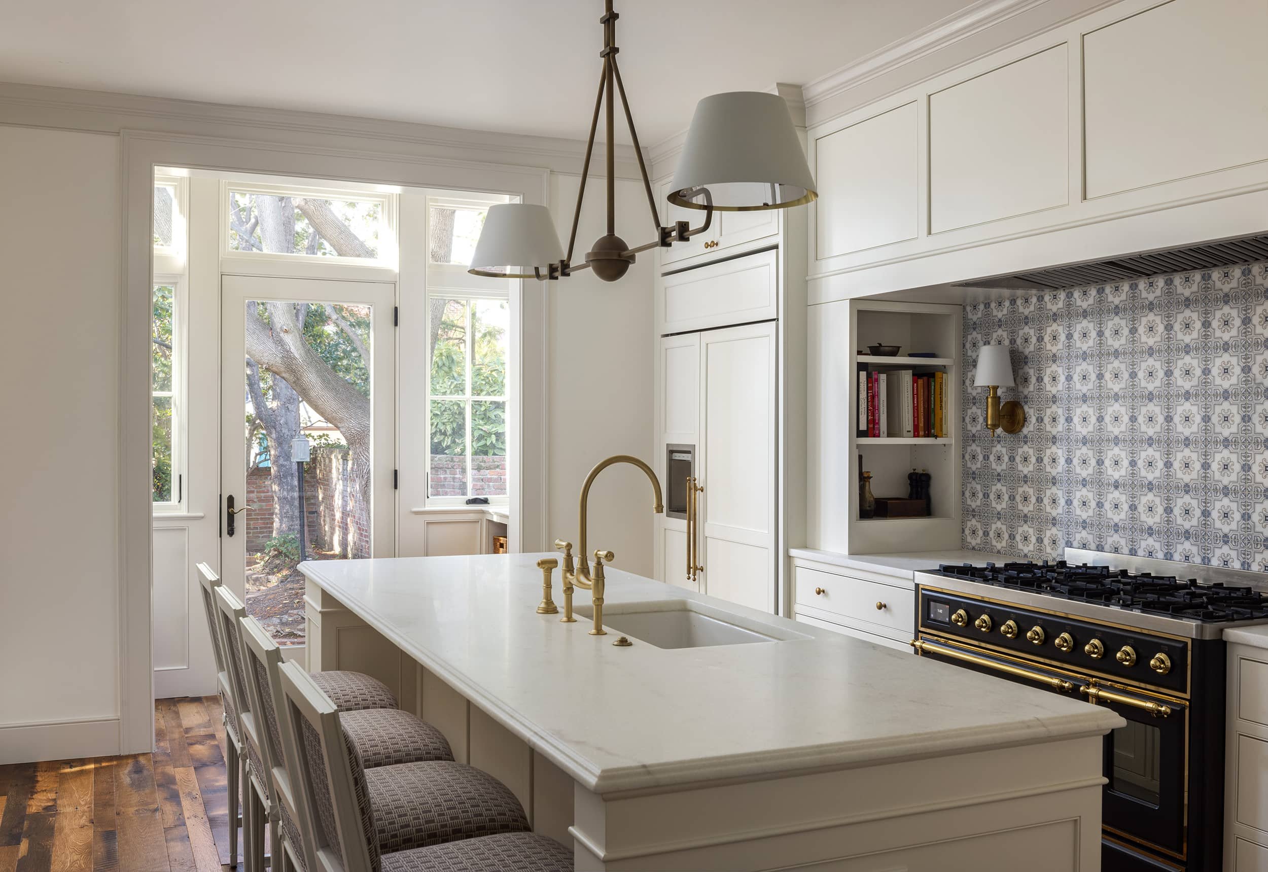 Bright kitchen with white cabinets, marble island, gold fixtures, patterned backsplash, built-in bookshelves, and a view of a backyard with trees through glass doors.