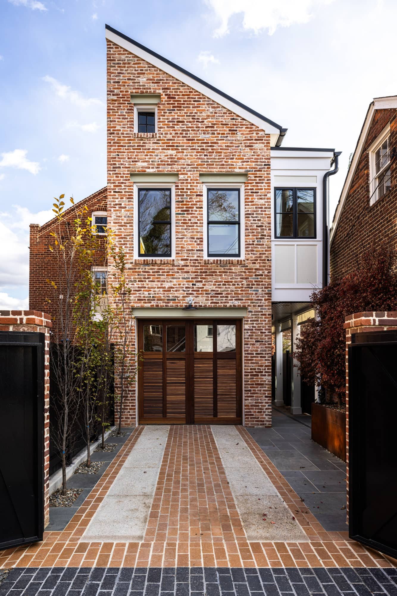 Brick house with multiple windows and a sloped roof. Wooden gate, and a brick-paved driveway.
