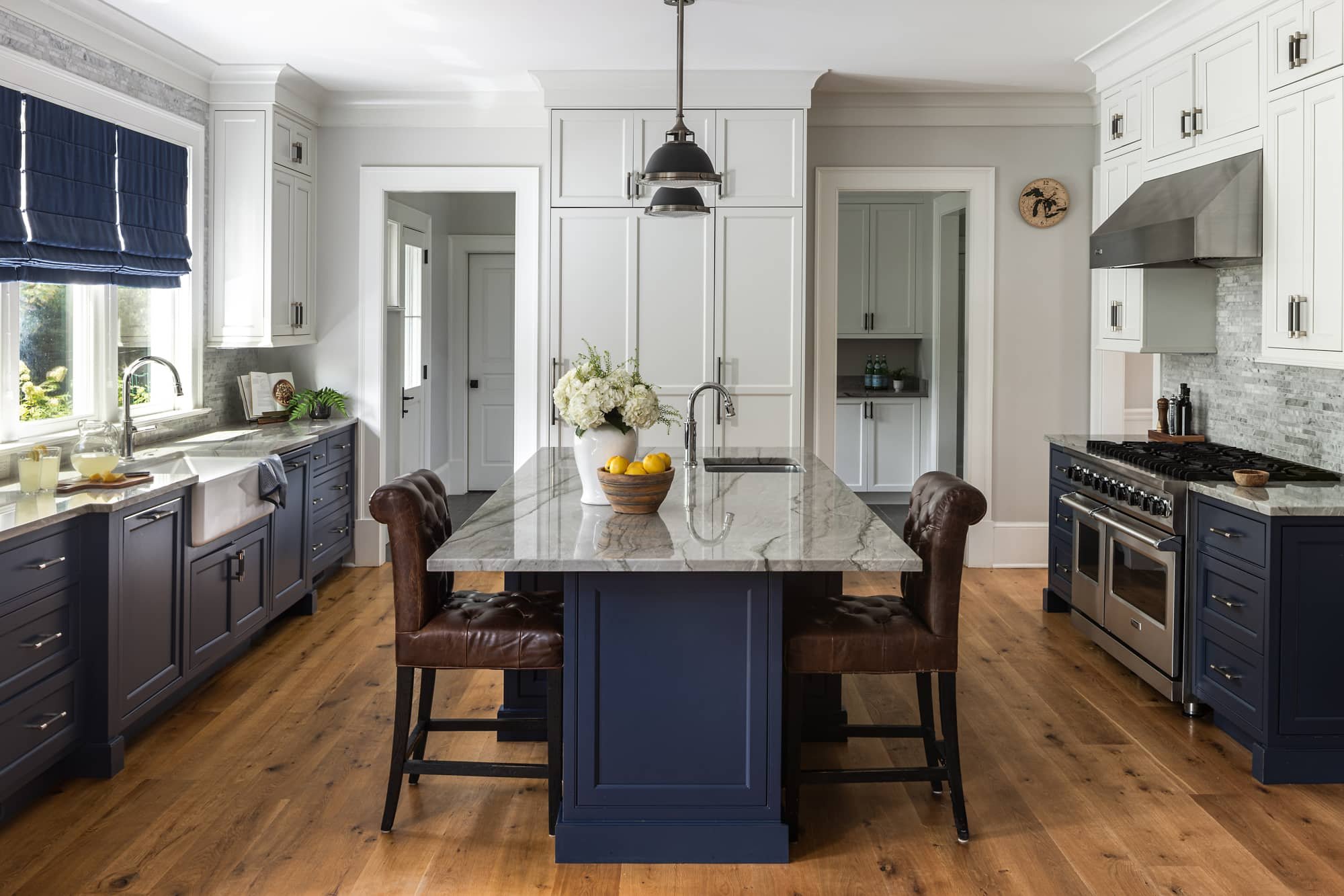 A spacious kitchen with white cabinets, blue lower cabinets, a gray marble island, hardwood floors, and a large window with navy curtains. There are brown leather chairs at the island, a vase with white flowers, and a bowl of lemons. Stainless steel appliances and a small shelf with bottles are also visible.