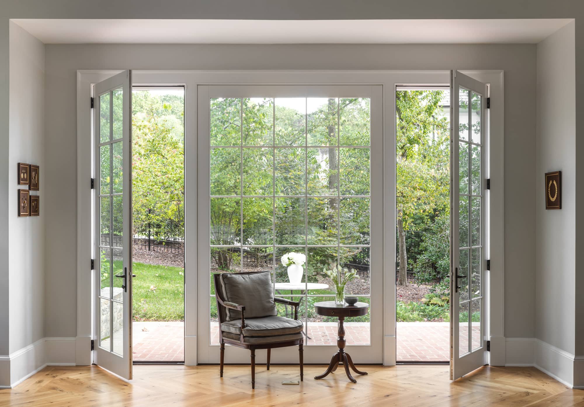 Living room with large glass door opening to a garden with trees and greenery, featuring a wooden chair with cushions, a small side table with a vase of white flowers, and artwork on the walls.