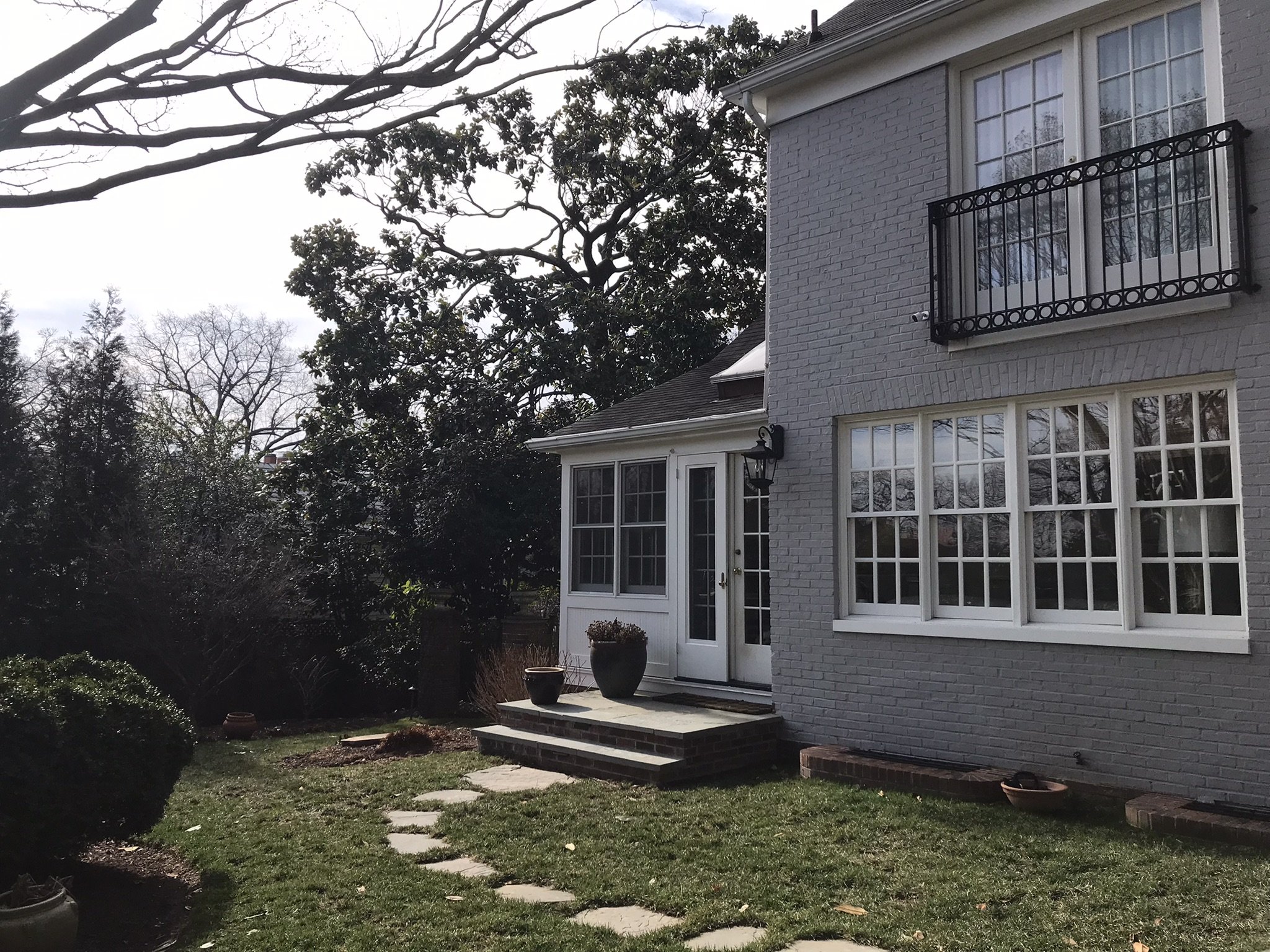 A view of the back of a gray brick house with a garden, patio steps, potted plants, a backyard with trees, and a cloudy sky.