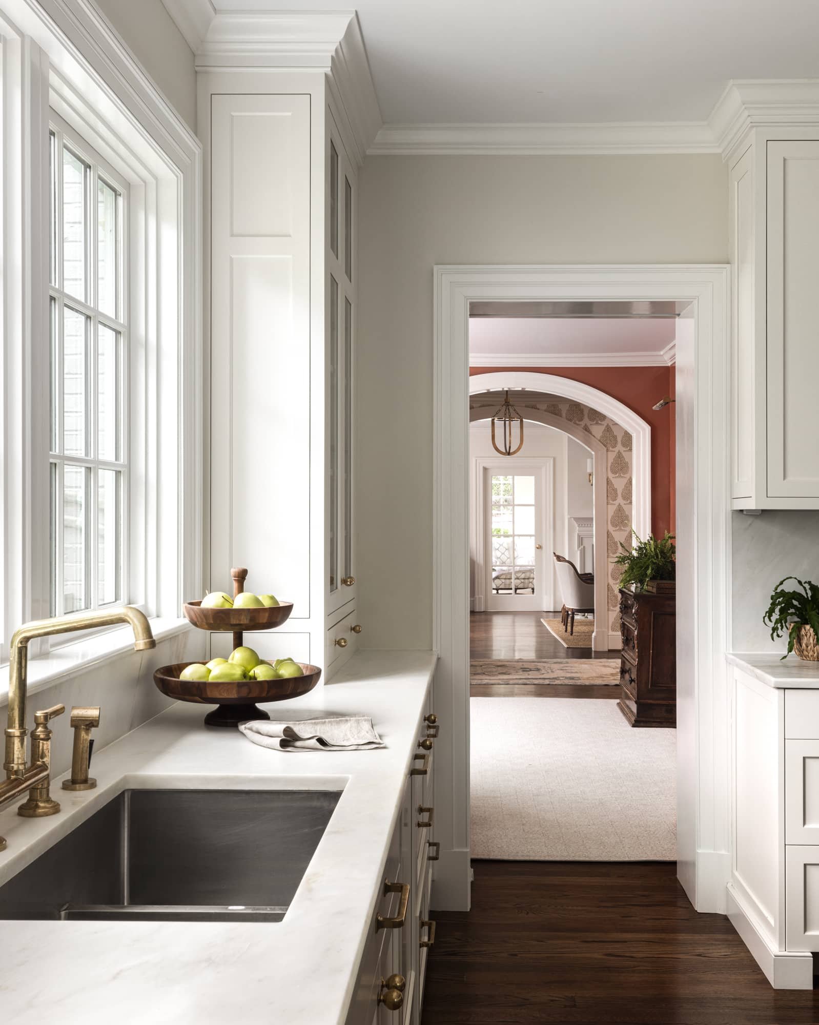 Bright kitchen with white cabinets, gold fixtures, and a marble countertop holding a bowl of green apples, overlooking a living room with a rug and sitting area.