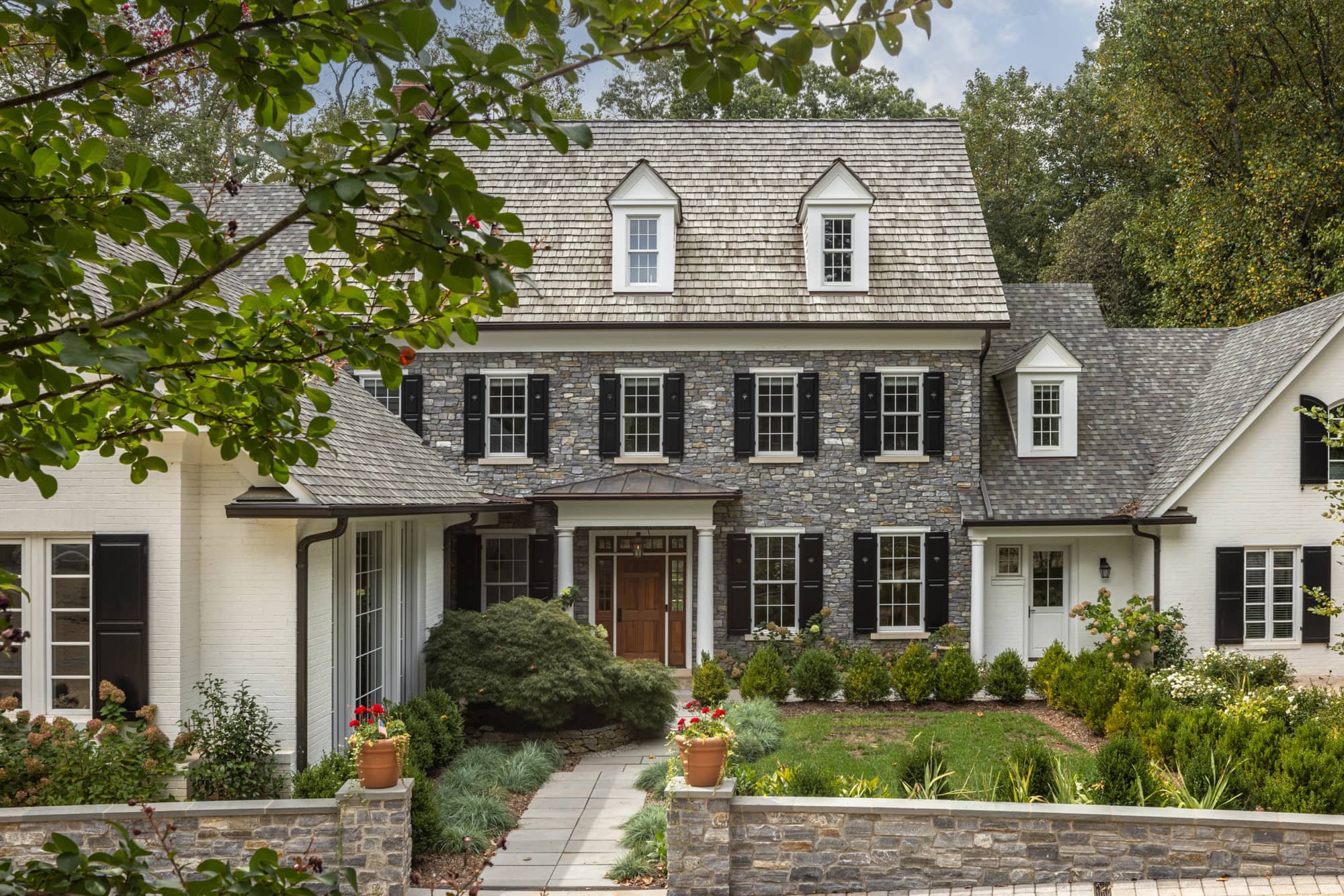 A two-story house with a stone facade and white walls, black shutters, and multiple windows. The front door is wooden with a small porch supported by white columns. The house has a gabled roof with small dormer windows and is surrounded by a landscaped garden with potted plants and bushes.