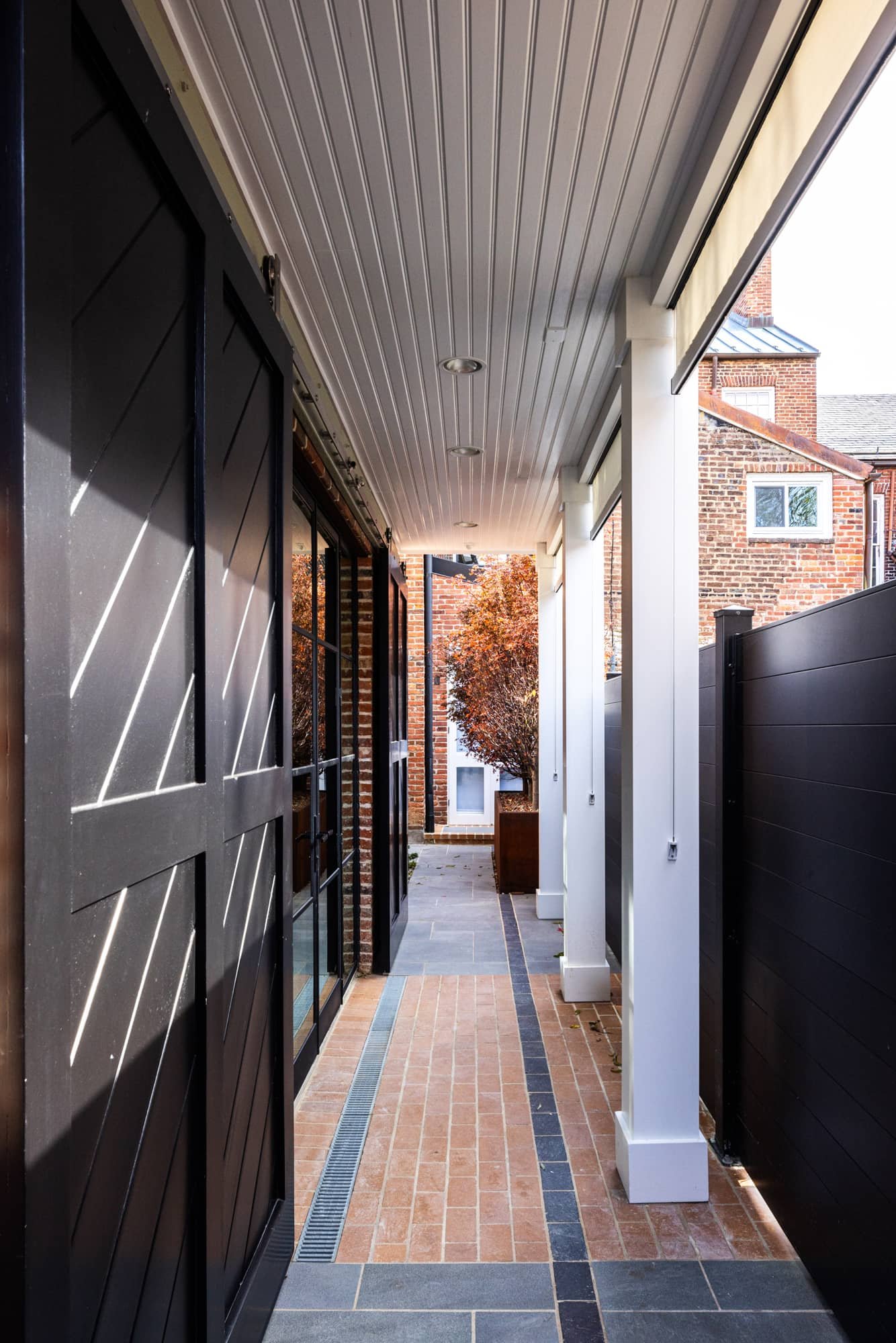 A narrow outdoor corridor of a modern building, with brick and black wall on the left and white columns on the right, leading to a small tree and a brick building in the distance.