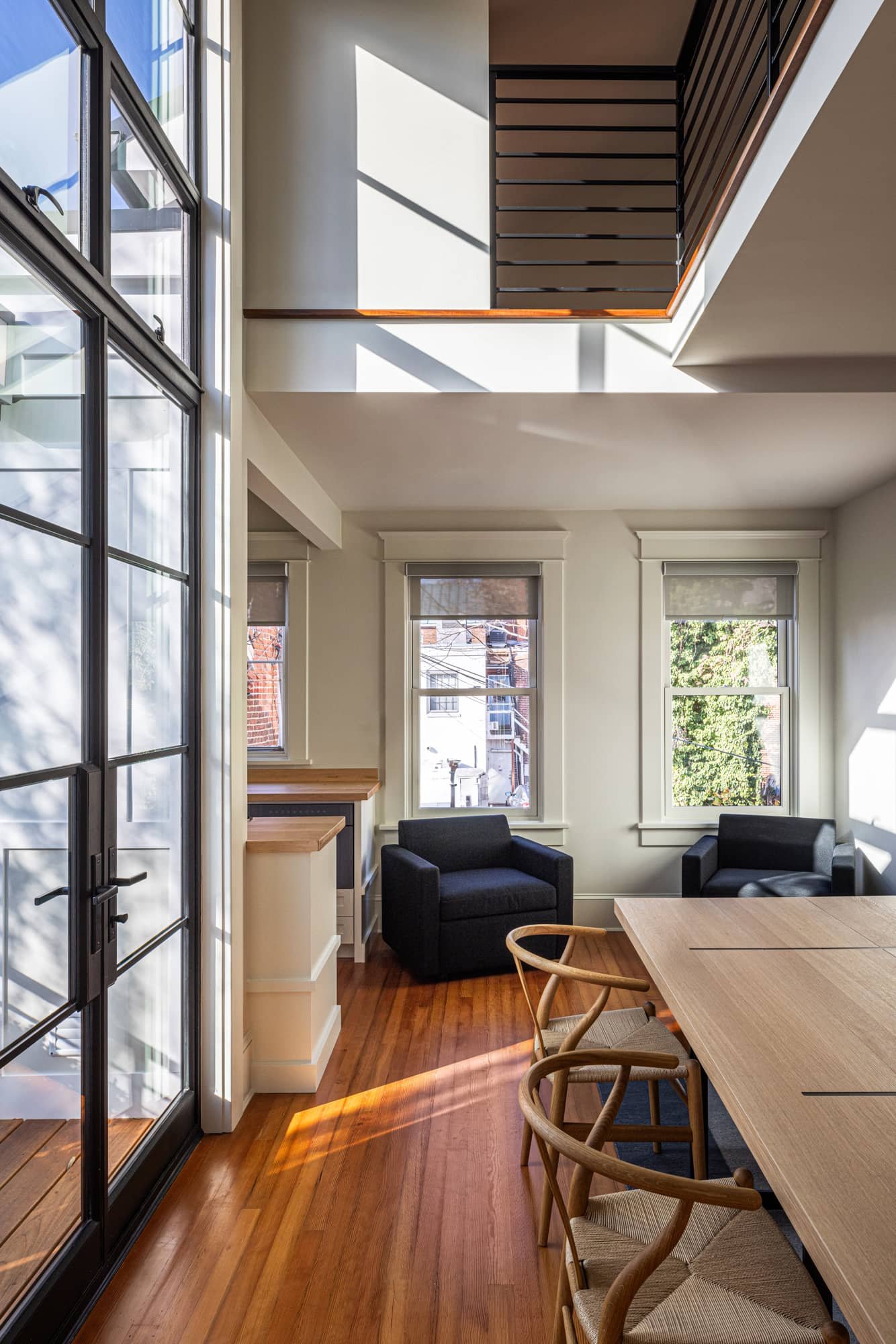 Bright living area with large windows, black armchairs, wooden chairs, and a wooden dining table, with sunlight streaming inside.