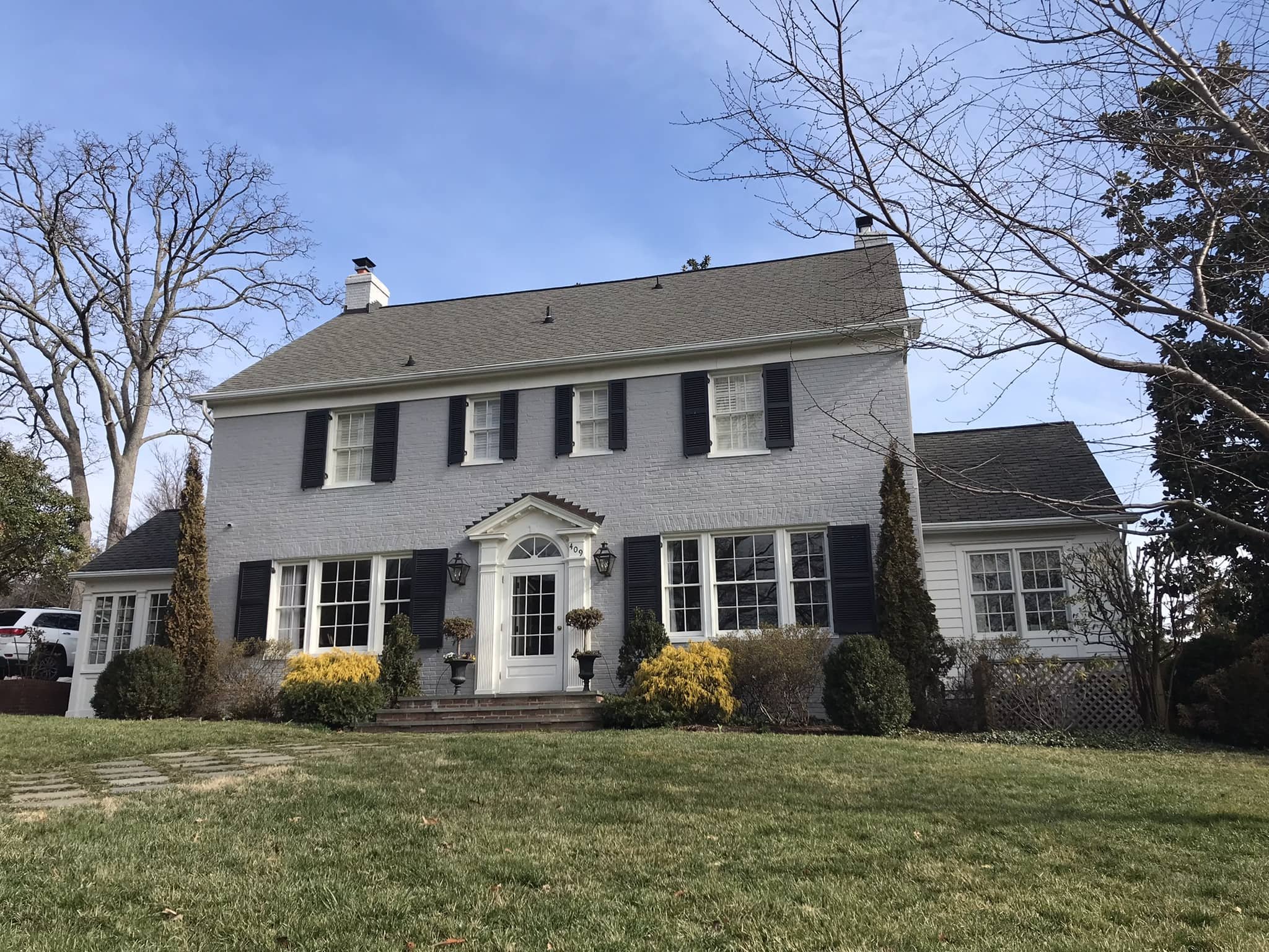 Front view of a two-story house with white brick and white siding, black shutters, a gray gabled roof, and a small set of brick steps leading to the front door. The house is surrounded by a lawn and trees, some with bare branches.