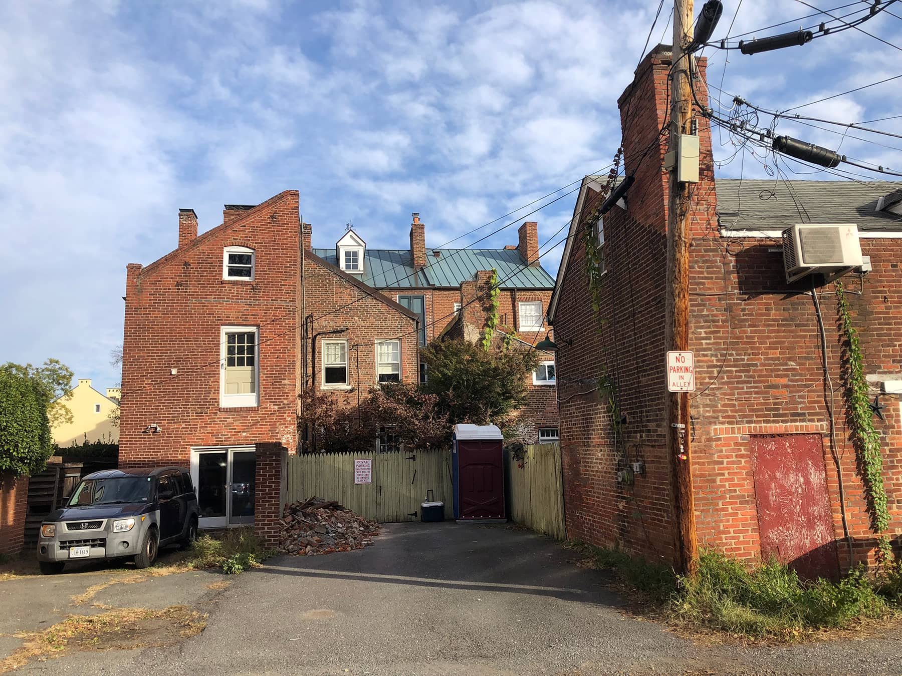 A small alleyway between brick buildings with parked cars, a portable toilet, a 'No Parking in Alley' sign, and a fenced area with trees and additional buildings in the background under a partly cloudy sky.