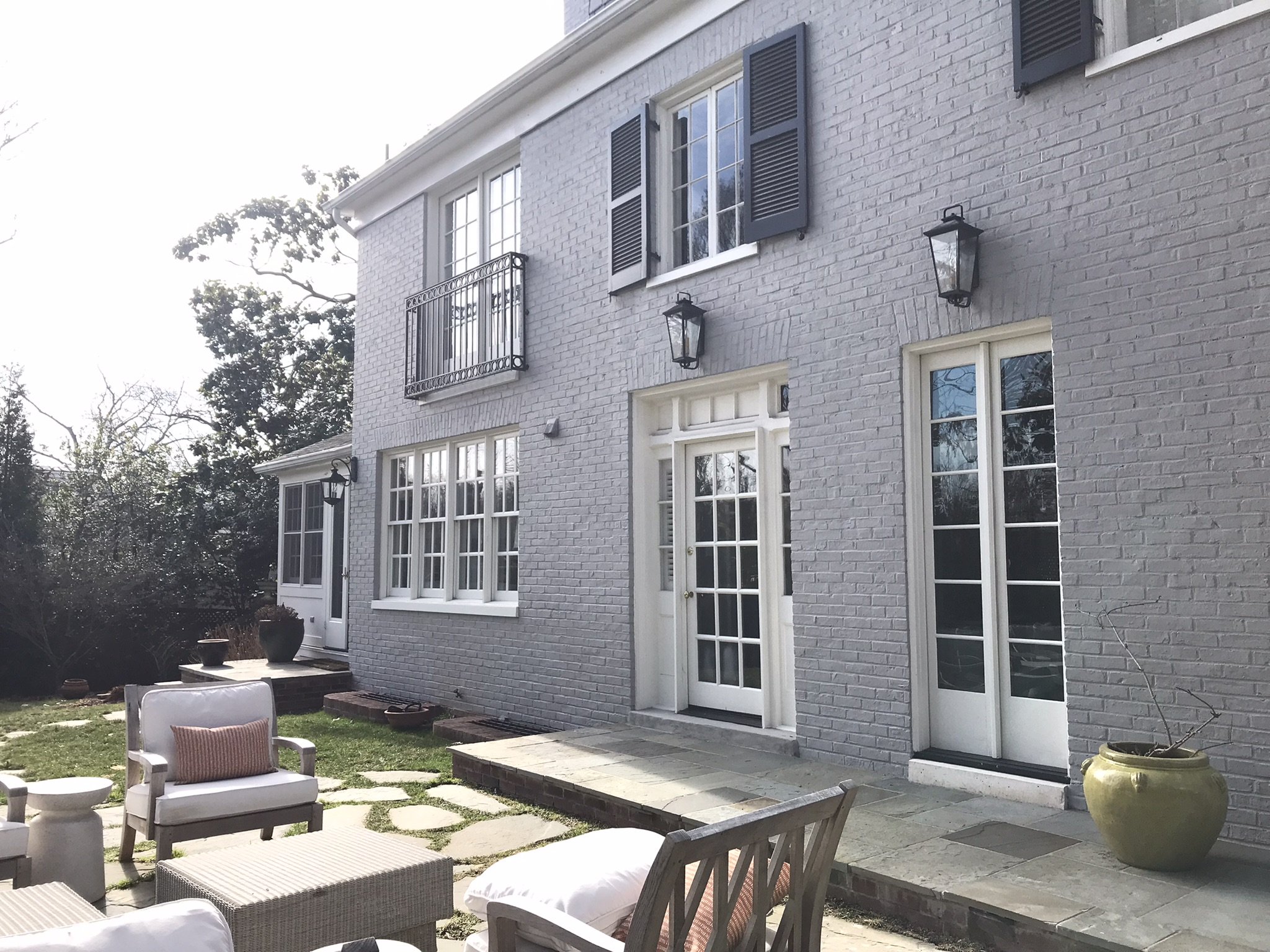 Backyard patio with white chairs, a small table, a large yellow pot, and a brick house wall with windows and black shutters.