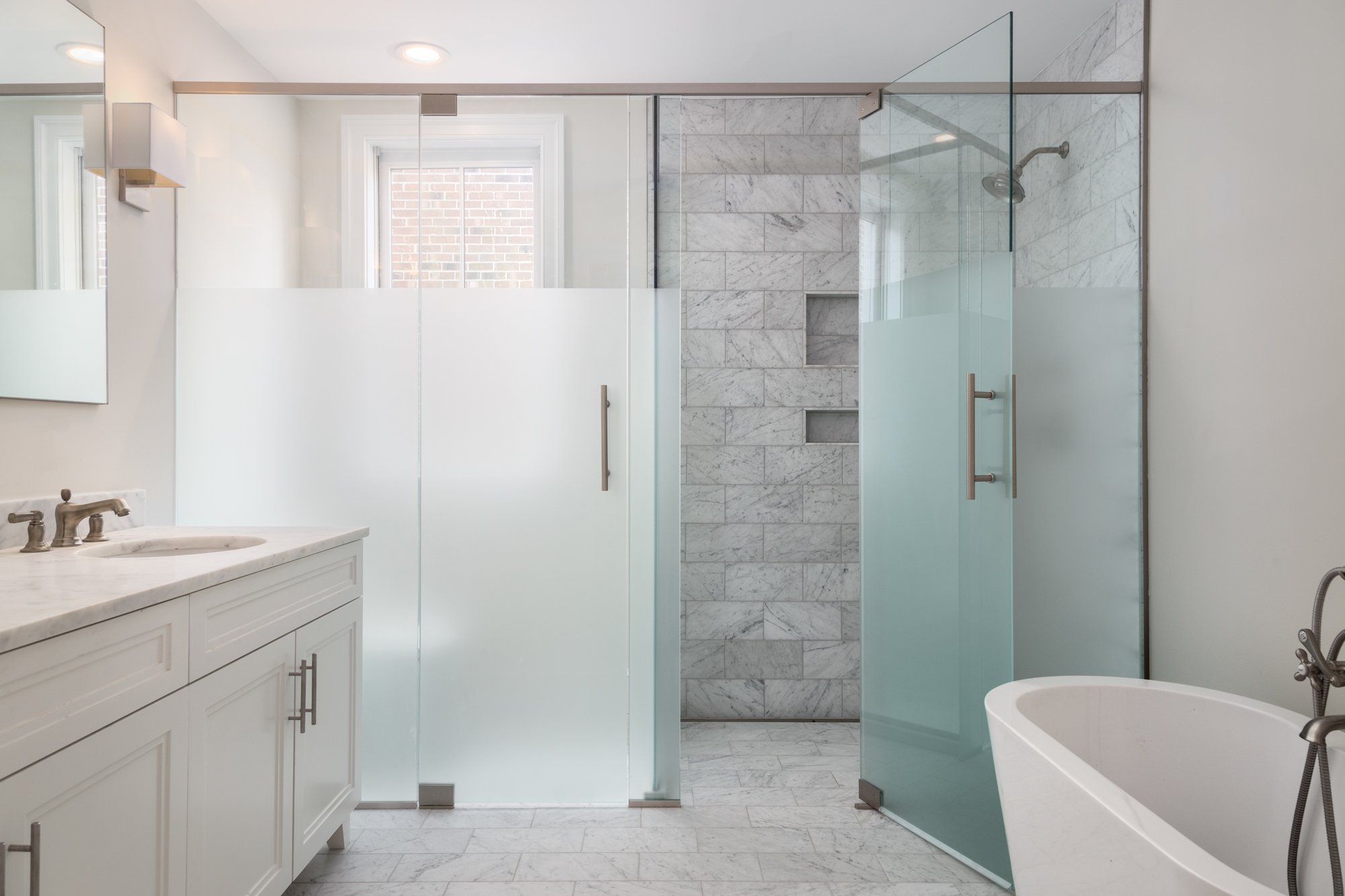 A modern bathroom with white cabinetry, a marble countertop, a glass shower enclosure with marble tiles, and a white freestanding bathtub.