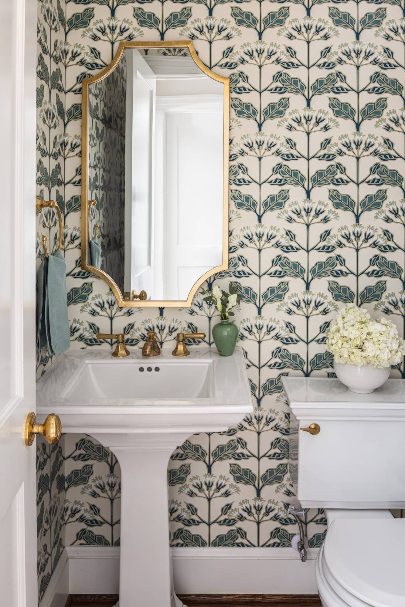A vintage-style bathroom with patterned wallpaper, a pedestal sink with brass fixtures, a gold-framed mirror, and a white ceramic vase with white flowers on the toilet tank.