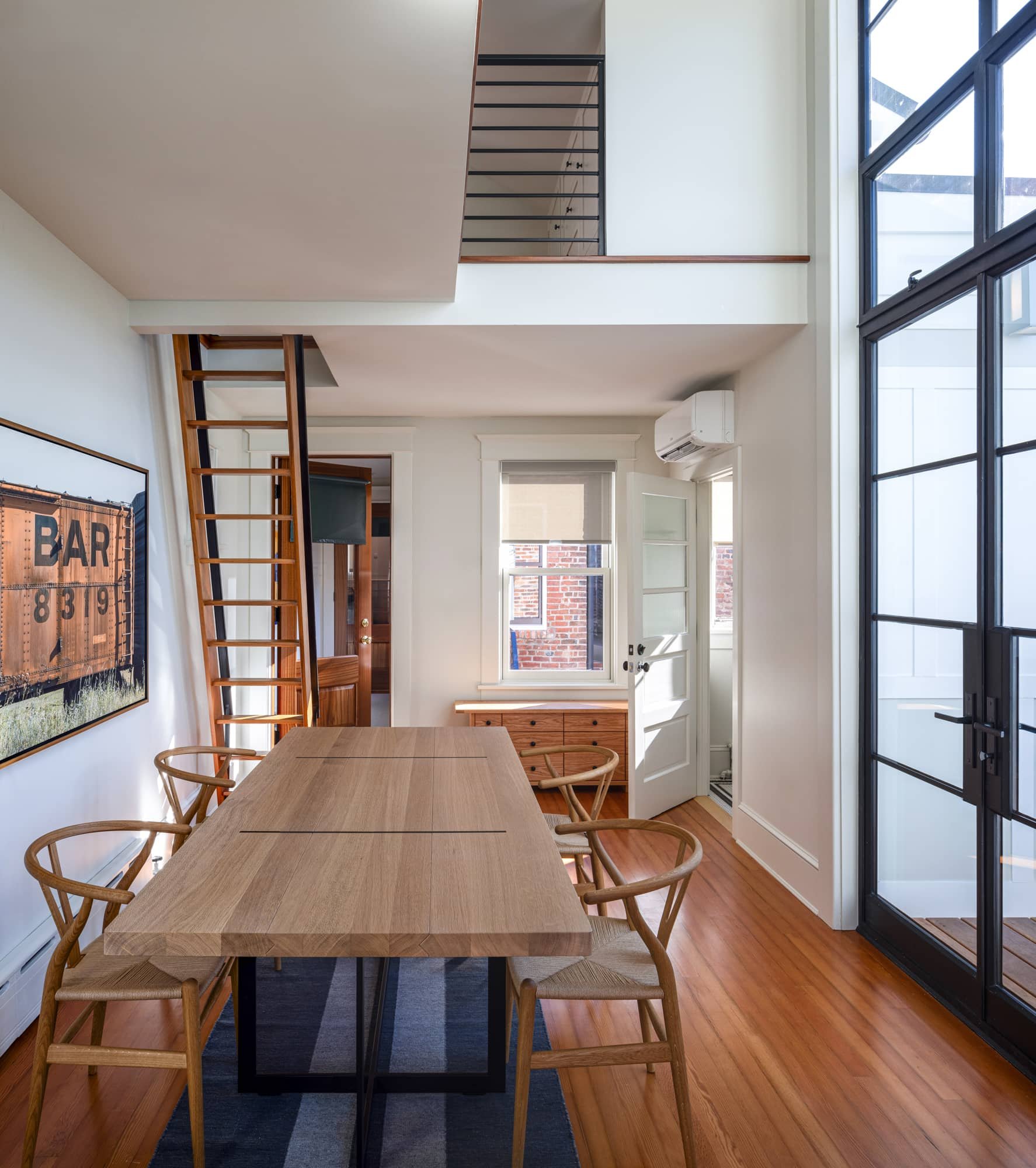 Interior of a modern dining area with a wooden table, six chairs, a black and white area rug, a white door, and large black-framed glass doors leading outside. There's a small window above a wooden sideboard, and a staircase with wooden steps and black railing going to an upper level.