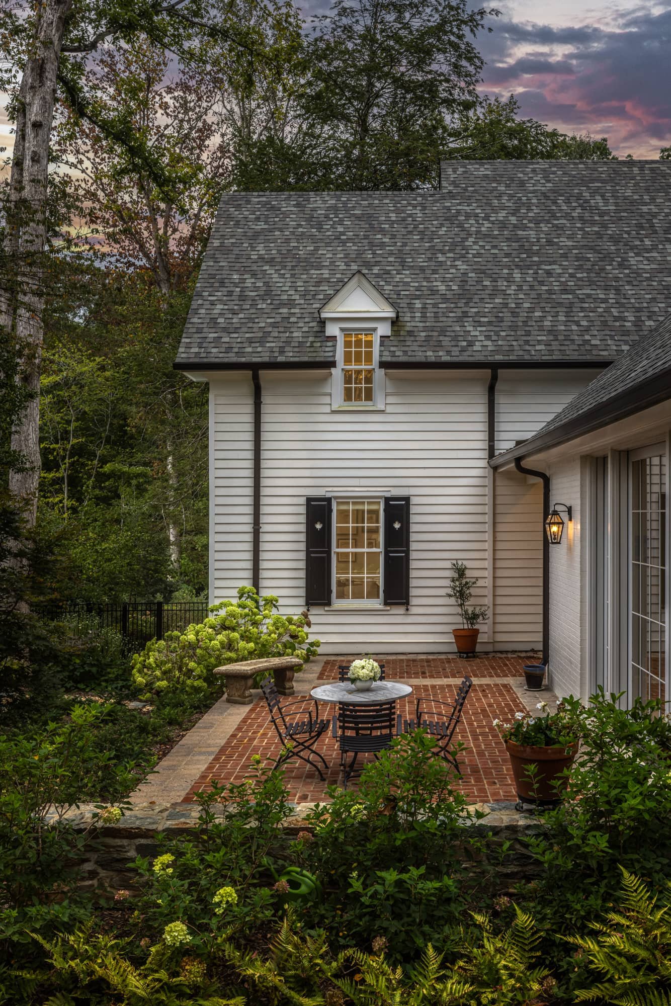 A cozy outdoor patio with a round table and four chairs, surrounded by lush green plants, on a brick surface next to a white house with black shutters, a lamp, and a small wooded backyard at dusk.