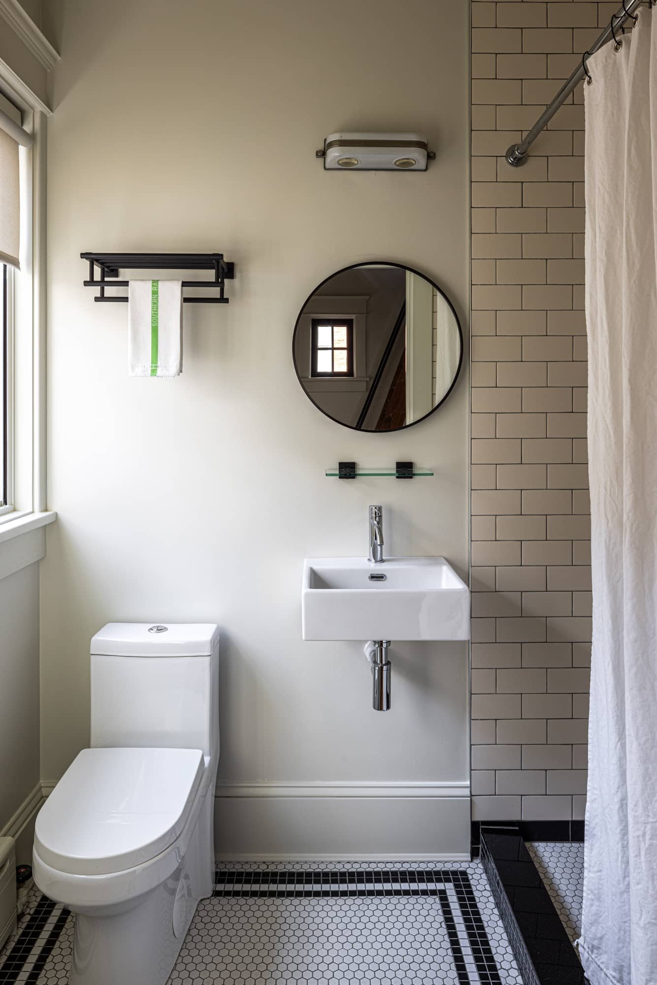 A small modern bathroom featuring a toilet, a wall-mounted sink, a round mirror, a black towel rack with a white towel, a window, a tiled shower curtain rod with a curtain, and a black and white mosaic tile floor.