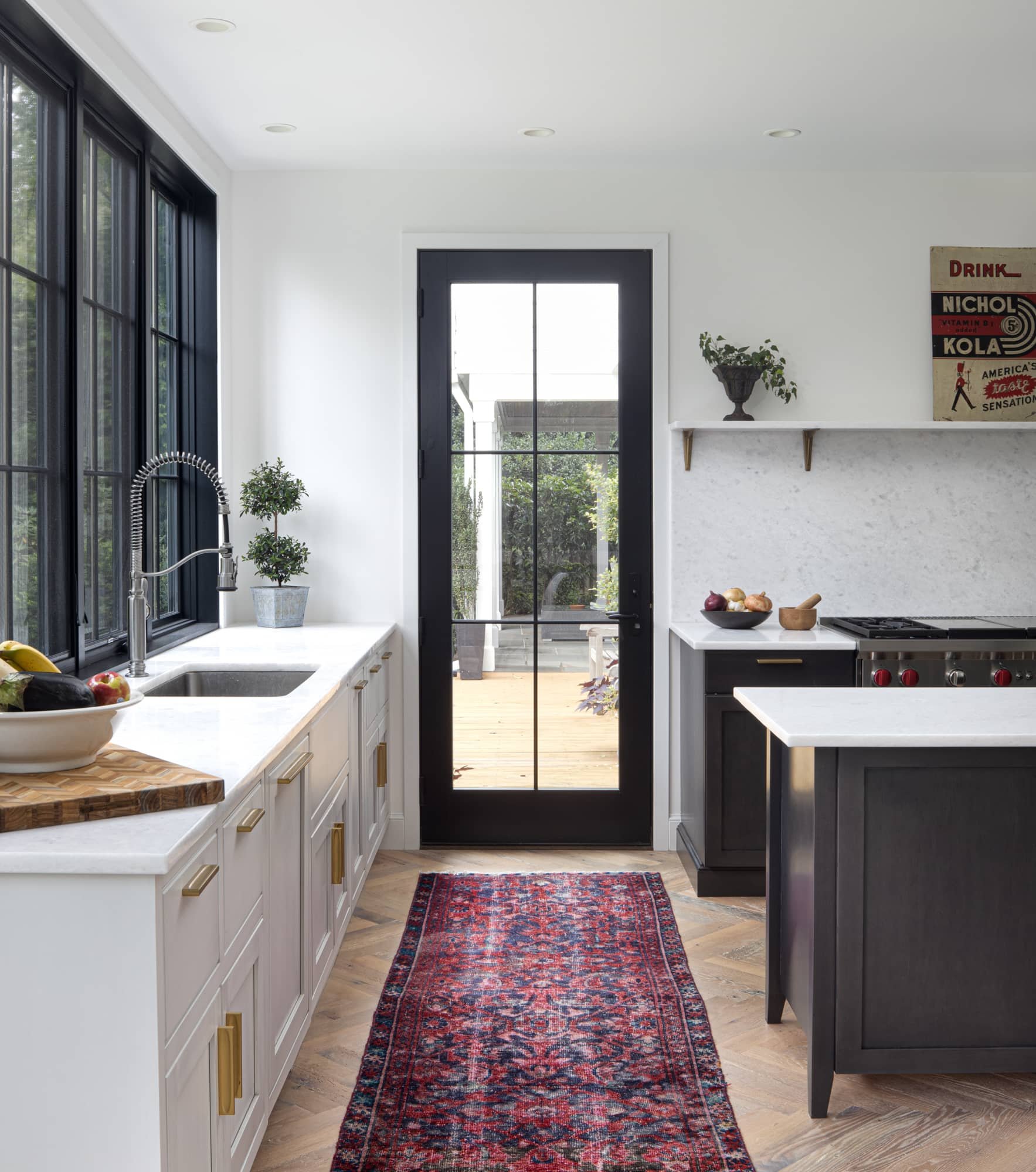 Modern kitchen with white and black cabinetry, a marble countertop, a sink under large black-framed windows, and a glass door leading to a deck outside. There are small potted plants, a bowl of fruit, a red patterned rug, and a piece of artwork on the wall.
