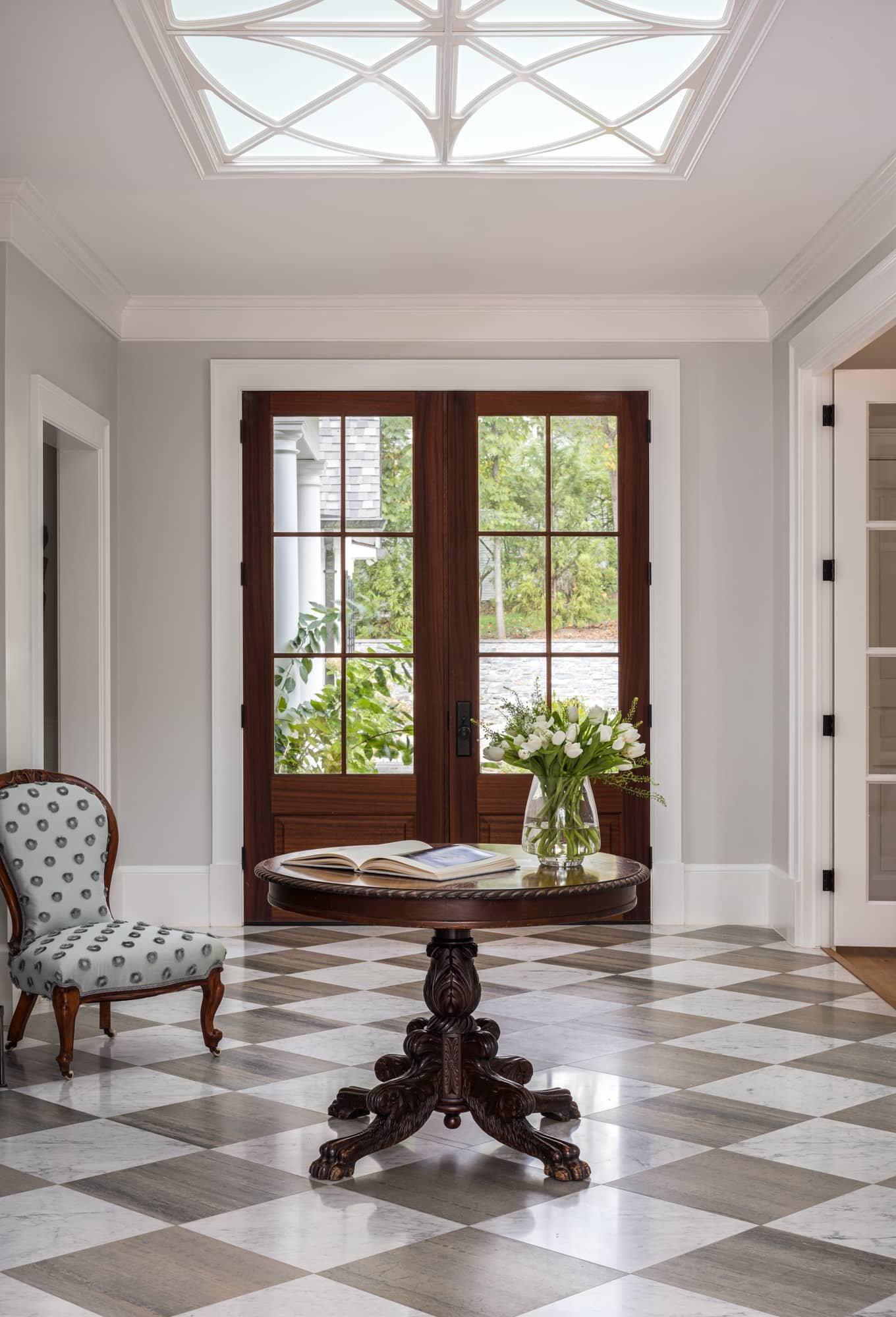 An interior room features a checkered floor, a round wooden table with a book and a glass vase of white flowers, a patterned armchair, and large glass doors with wooden frames leading outside, with greenery visible beyond.