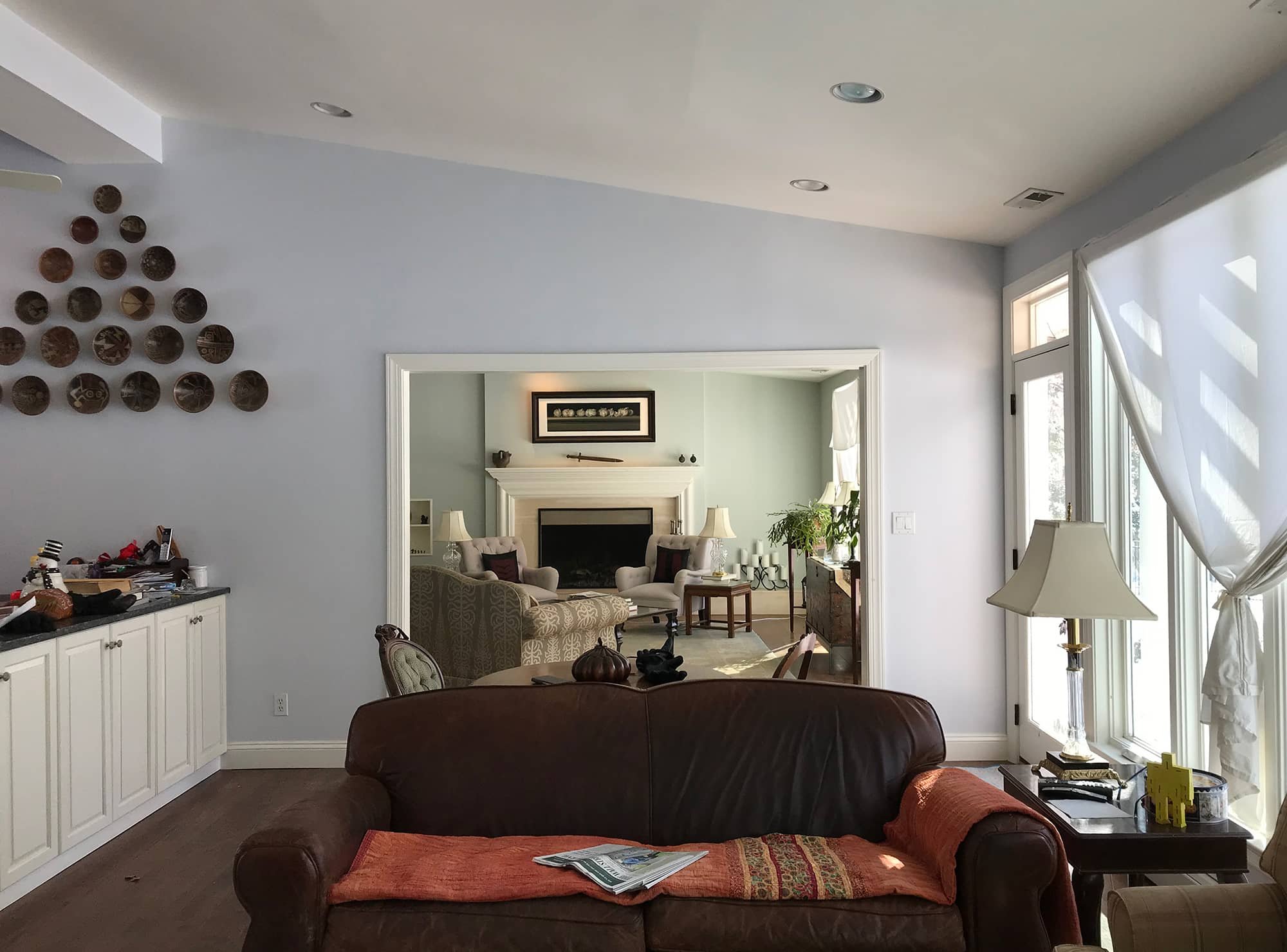 Living room with white cabinets, a leather sofa with an orange blanket, and a side table with a lamp; leads to another room with armchairs and a fireplace.