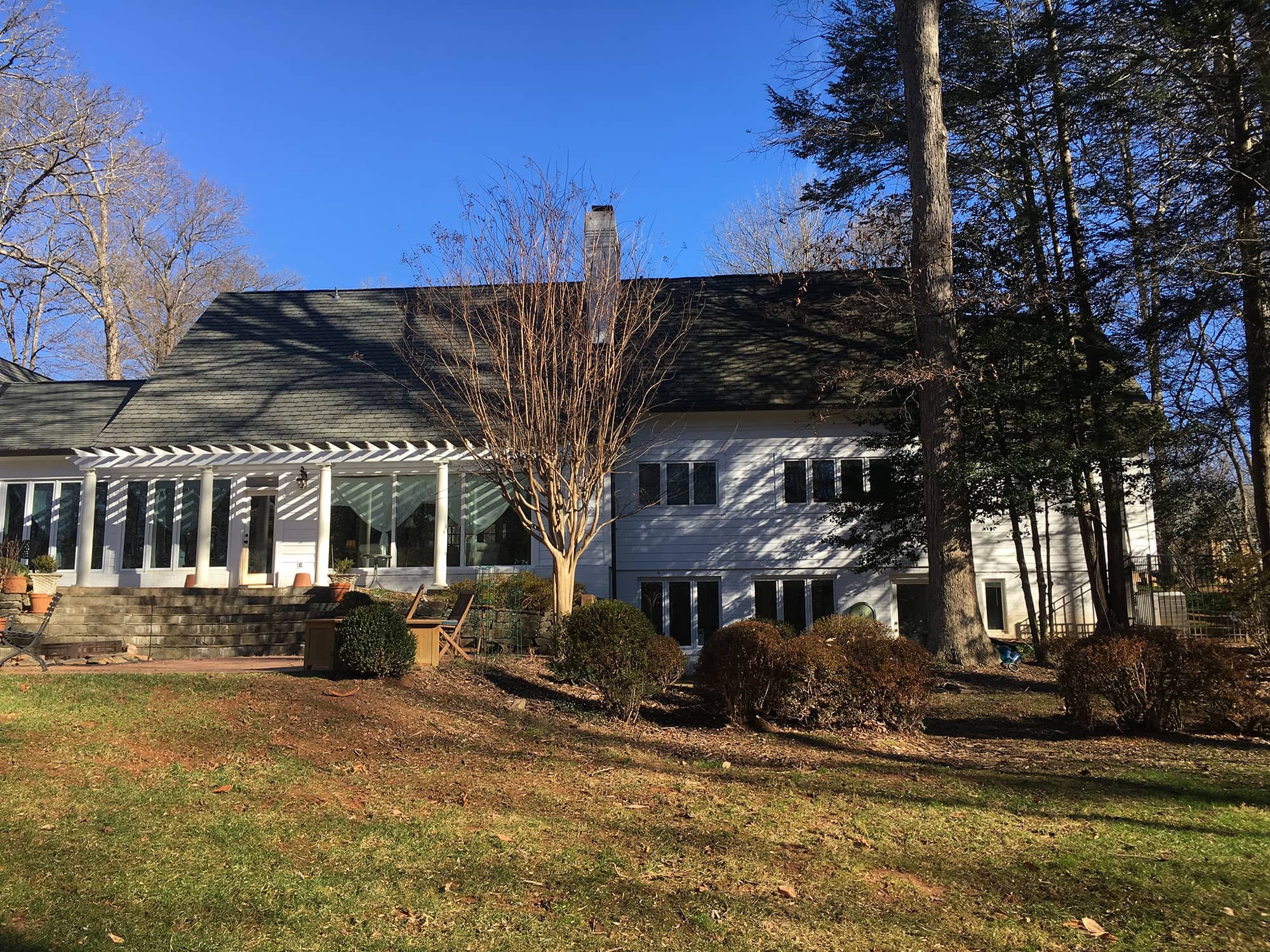 A large house with white exterior walls, black roof, and a screened porch with white pillars. The house is surrounded by trees with some leaves fallen, shrubs, and a grassy yard, under a blue sky.