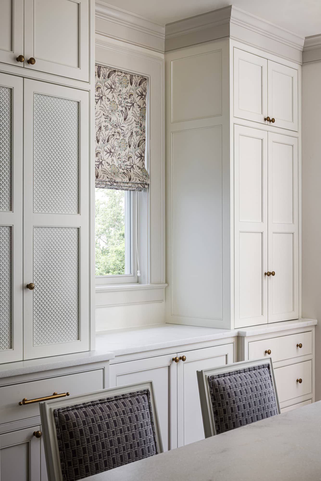 White kitchen cabinets and a window with a floral Roman shade, part of a dining area with chairs.