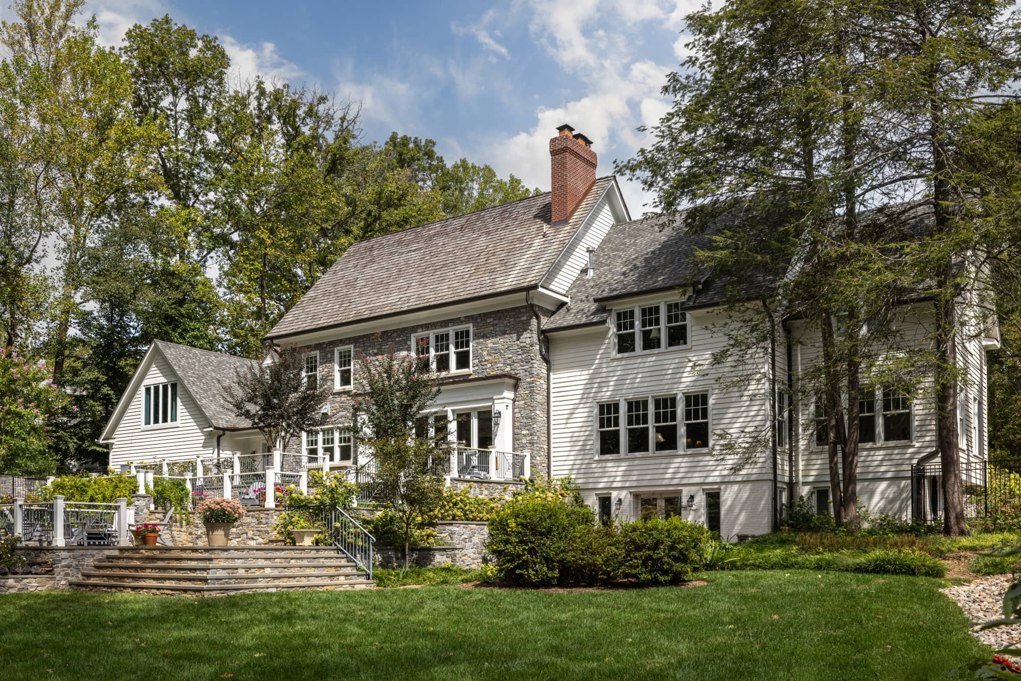 Large white house with stone accents, multiple windows, and a chimney, surrounded by trees and a manicured lawn with steps and a patio area.