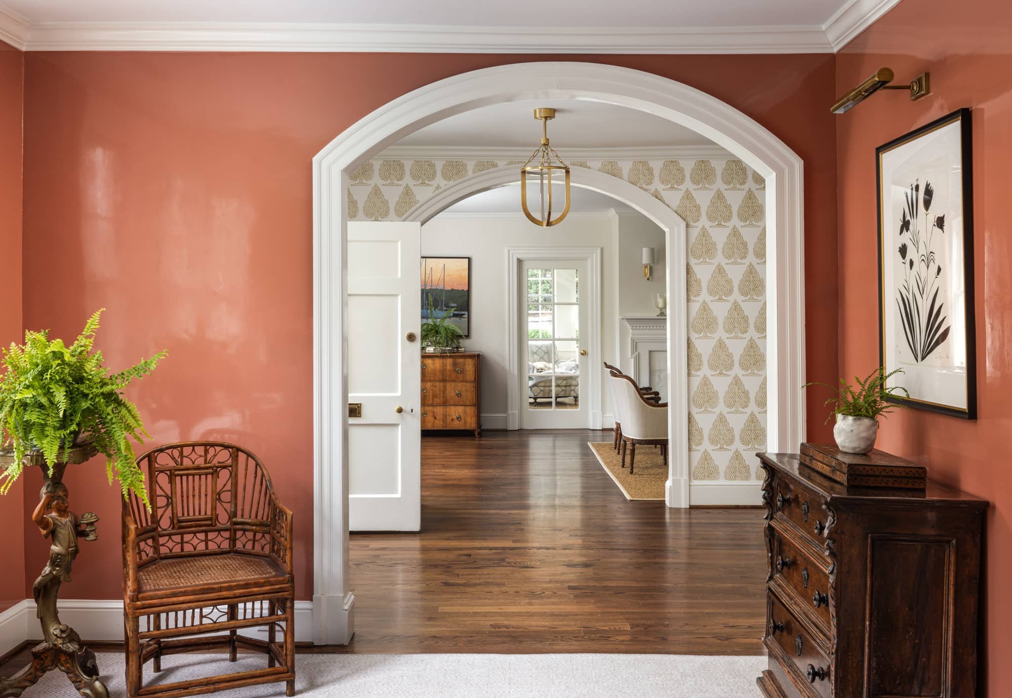 Interior view of a home with a front door, terracotta-colored walls, white trim, and a wooden floor. There is a vintage wooden chest of drawers, a potted fern andart piece. Multiple archways lead to a sunlit room with wallpapered walls.