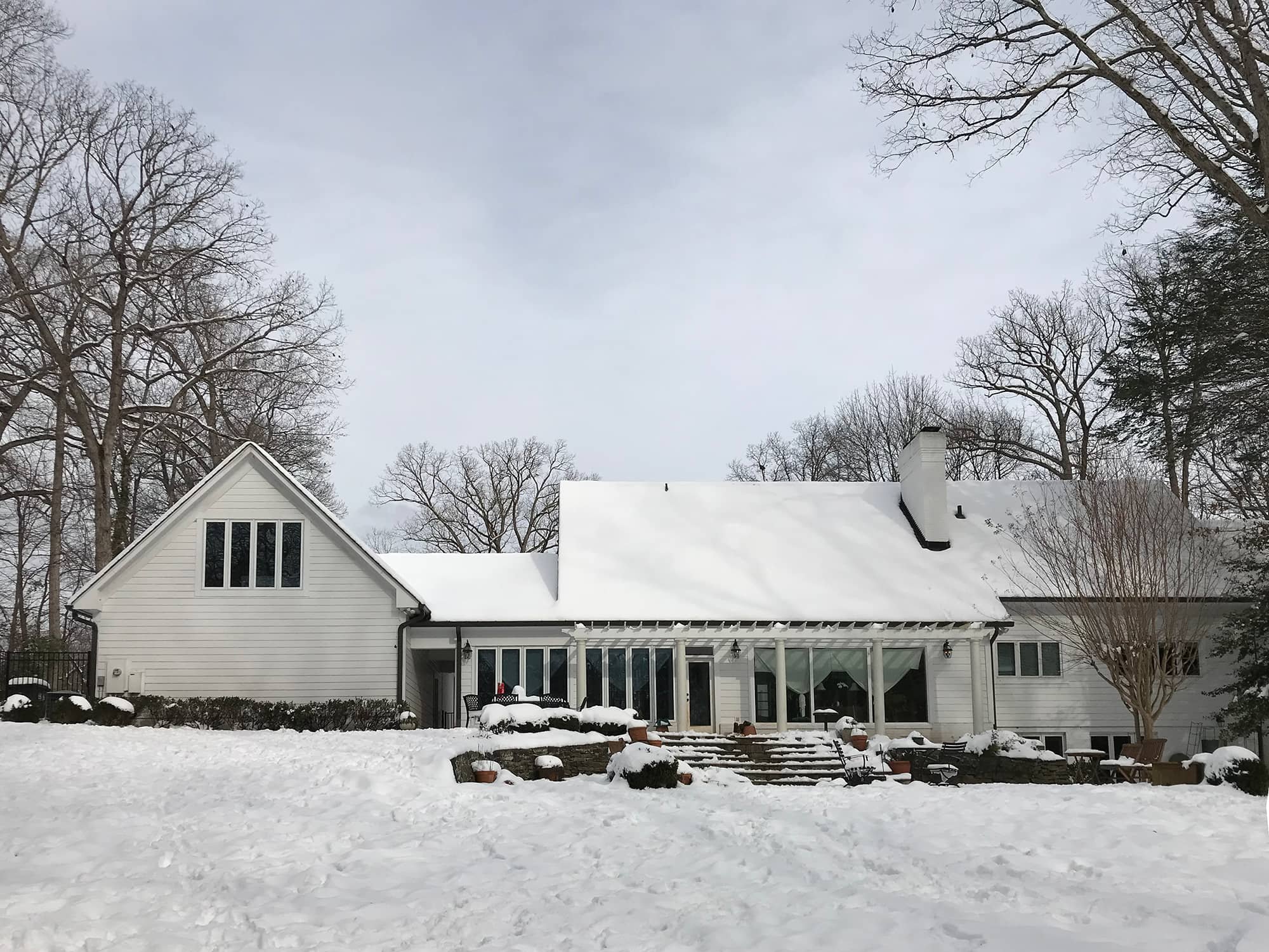 Snow-covered house with white siding, large windows, and a porch, surrounded by leafless trees and snow-covered ground.