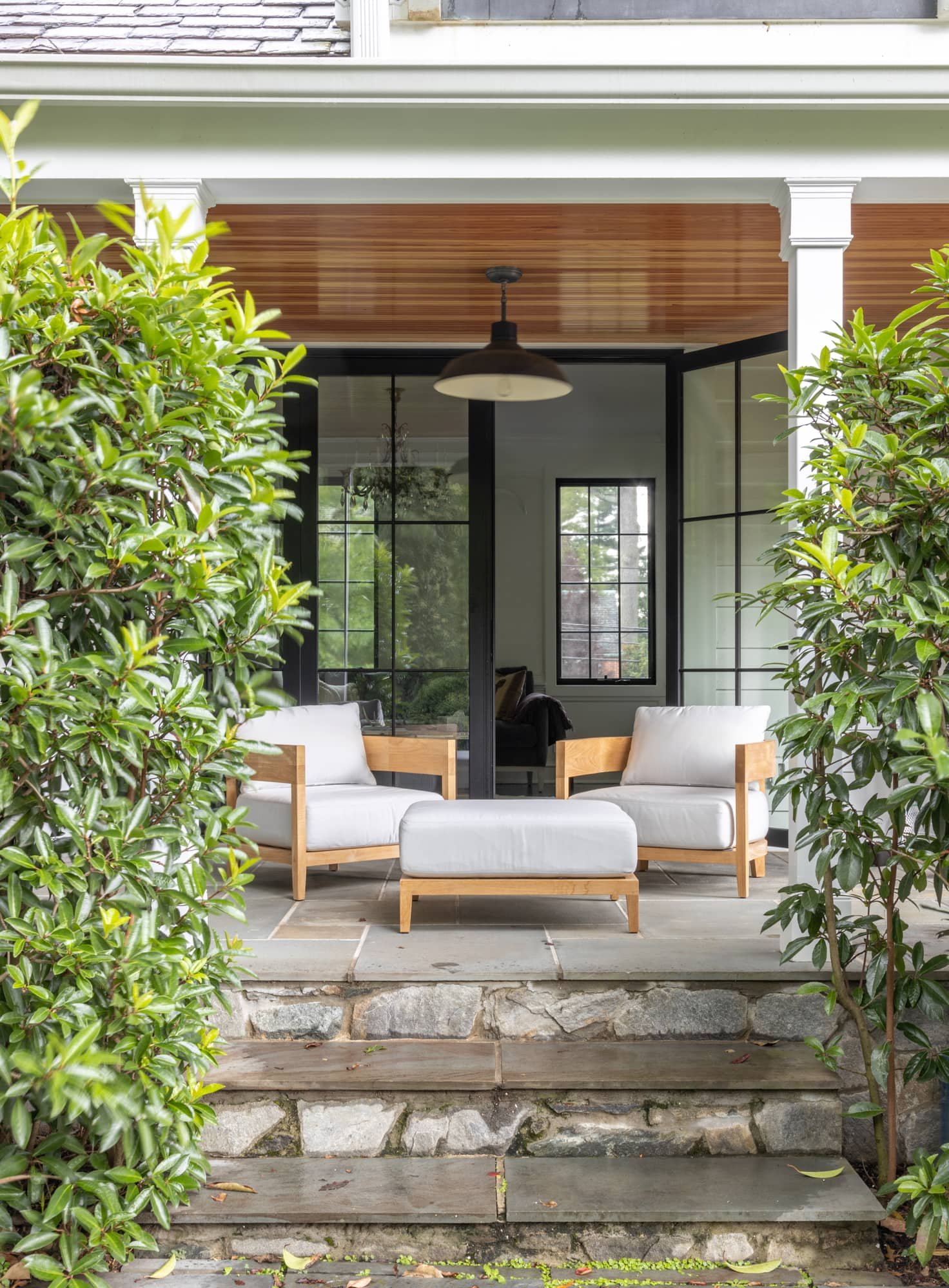 A front porch with two white cushioned armchairs and a matching ottoman, surrounded by greenery with stone steps leading up to the porch.