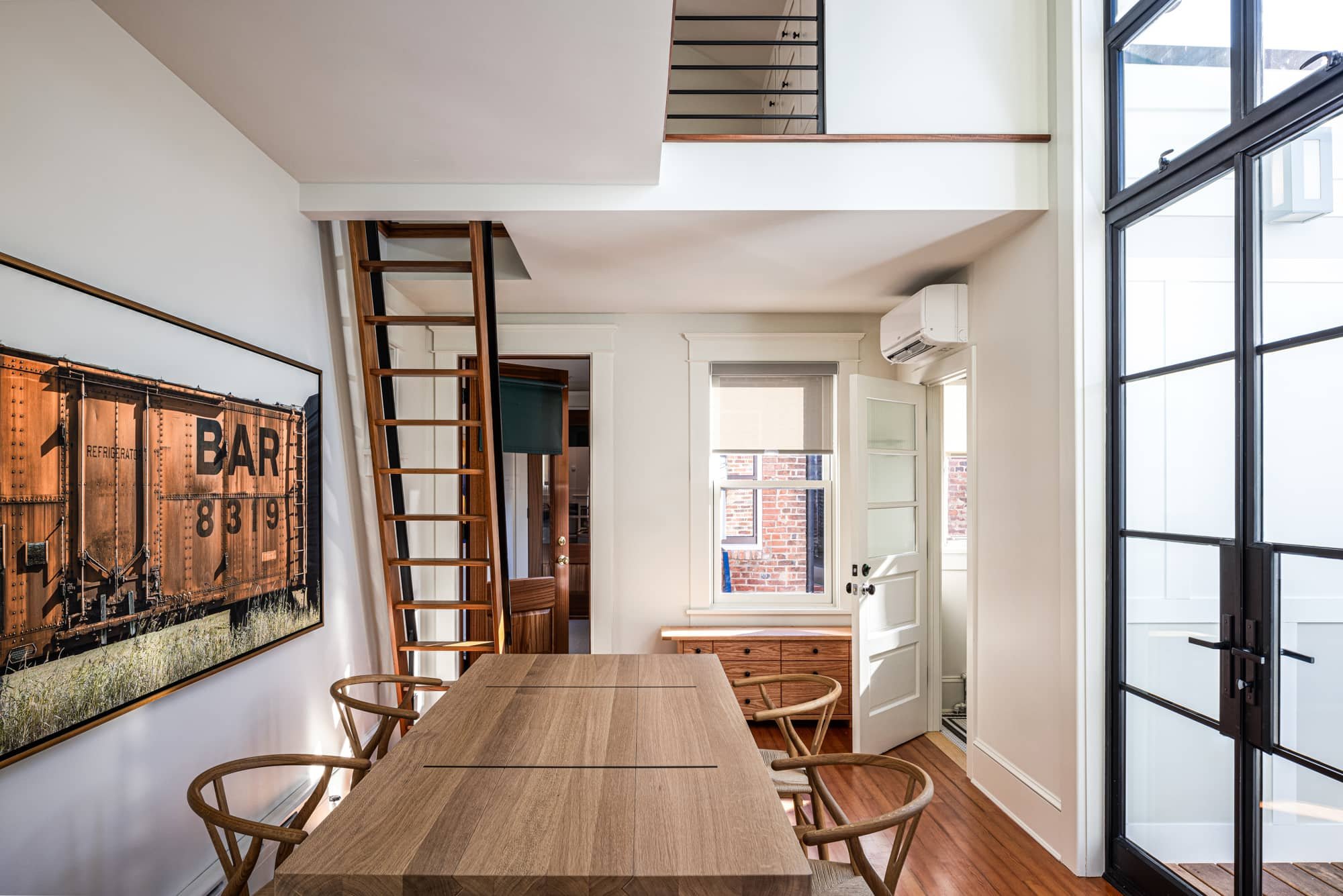 Interior of a modern dining room with a wooden table, chairs, a large wall art featuring a train car, a window with a view of a brick building, a door, a small wooden cabinet, and black-framed glass door leading outside.