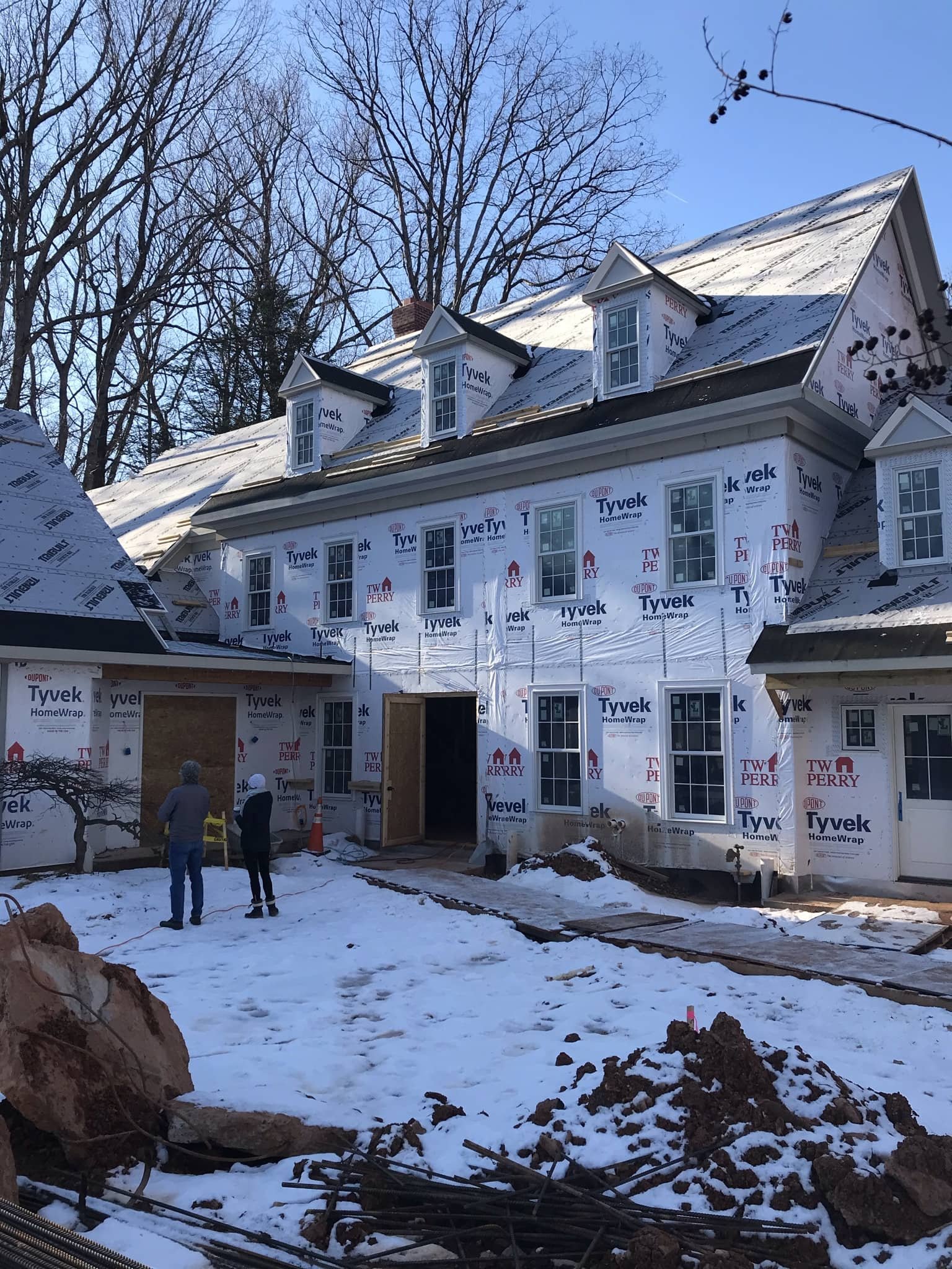 A house under construction with a snowy yard and partly finished exterior wall wrapped in Tyvek weatherproofing material. Two workers are present on the site, and there are trees and a clear blue sky in the background.