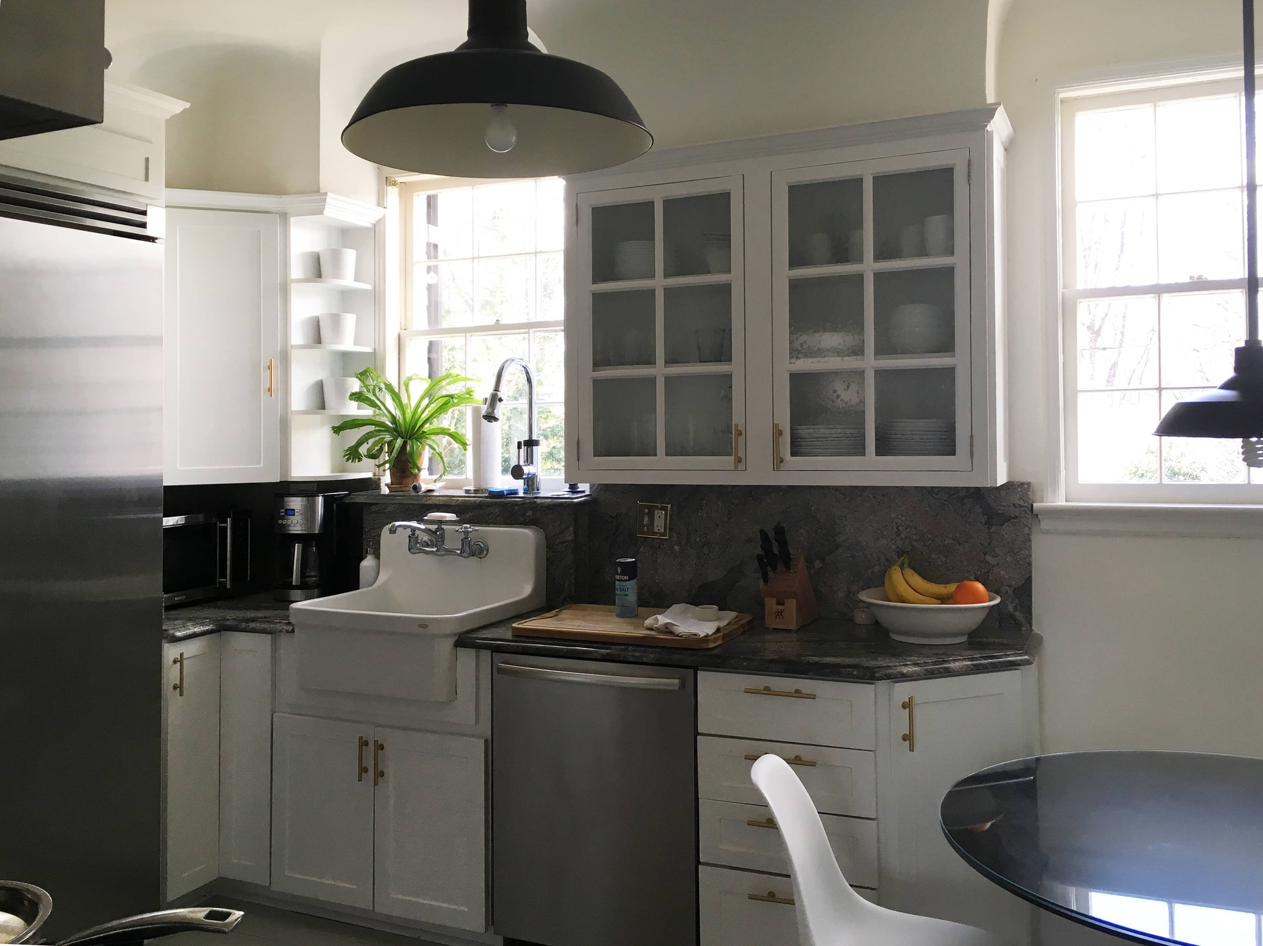 A bright kitchen with white cabinets, a black and gray marble countertop, a farmhouse sink, and large windows. There are some bananas and an orange in a white bowl on the counter, and a potted plant near the window.