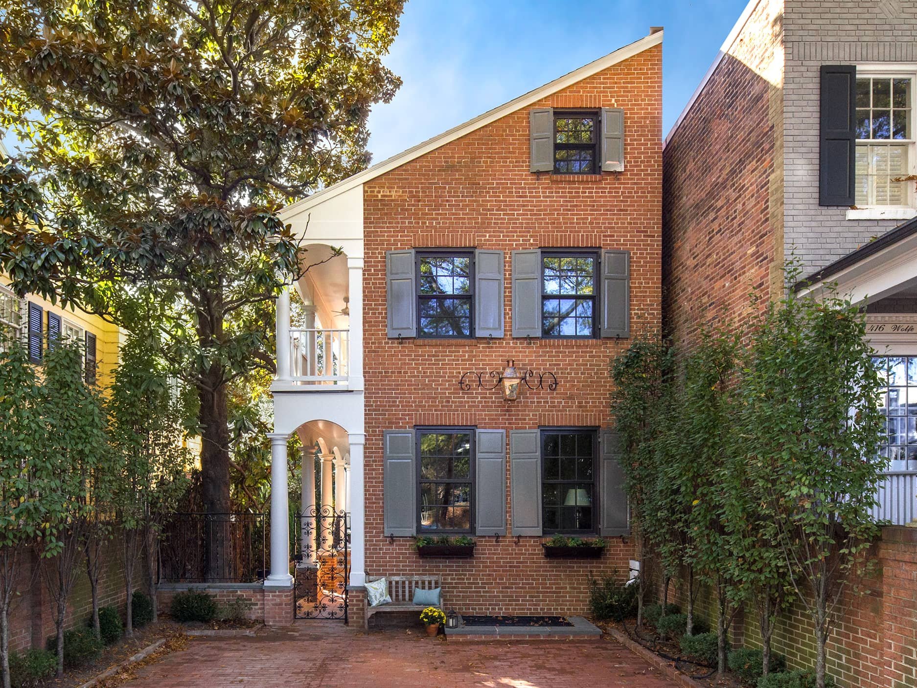 Front view of a three-story brick townhouse with gray shutters and black window frames, surrounded by trees and a brick-paved driveway.