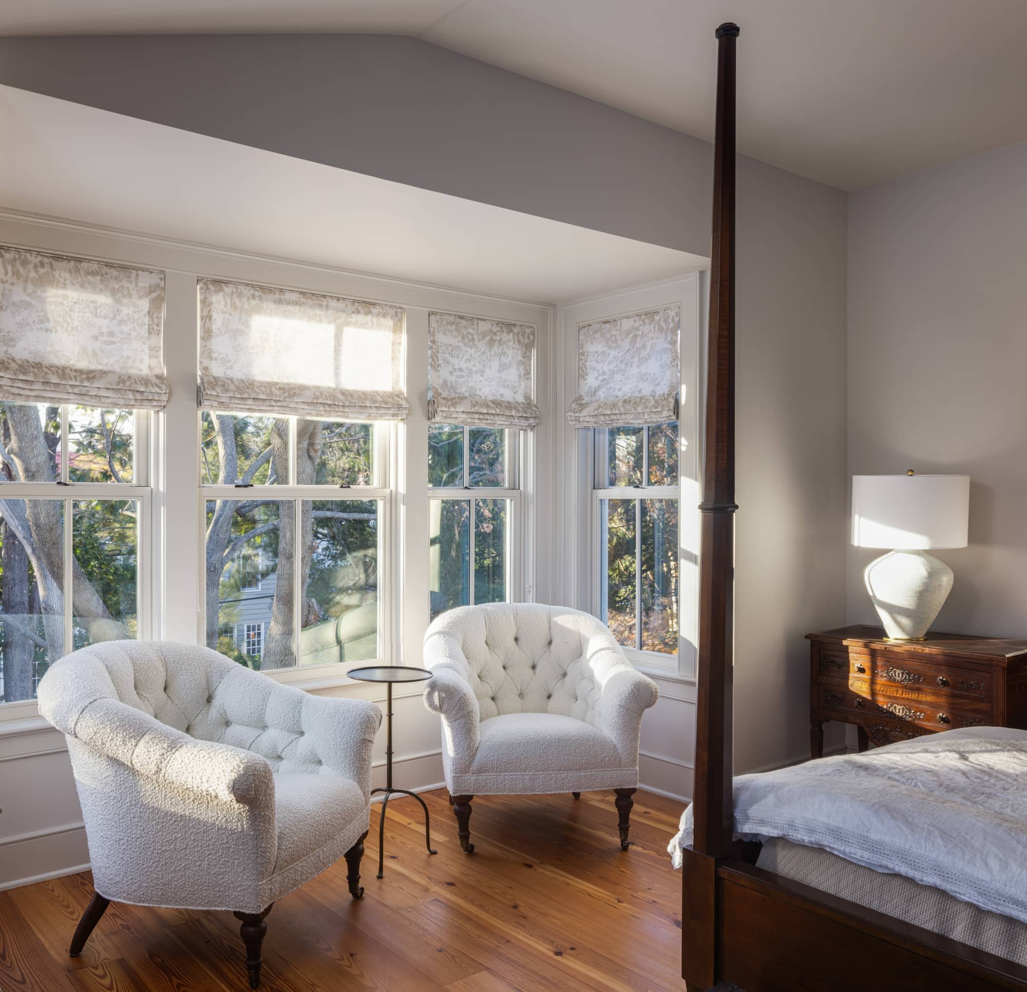 A bedroom with a bed, nightstand, lamp, and two white upholstered chairs near a bay window with white curtains, wooden floors, and natural light.