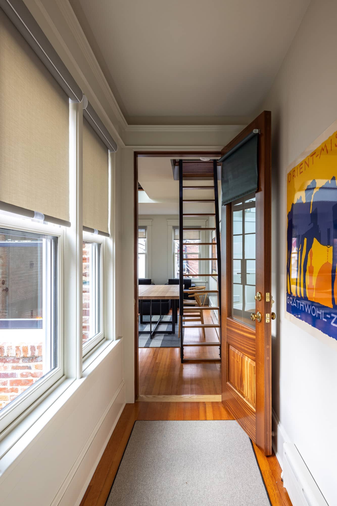 View from a cozy entrance hallway with wooden flooring, a closed wooden door with glass panels, and a colorful abstract poster on the wall, leading into a dining area with a ladder.