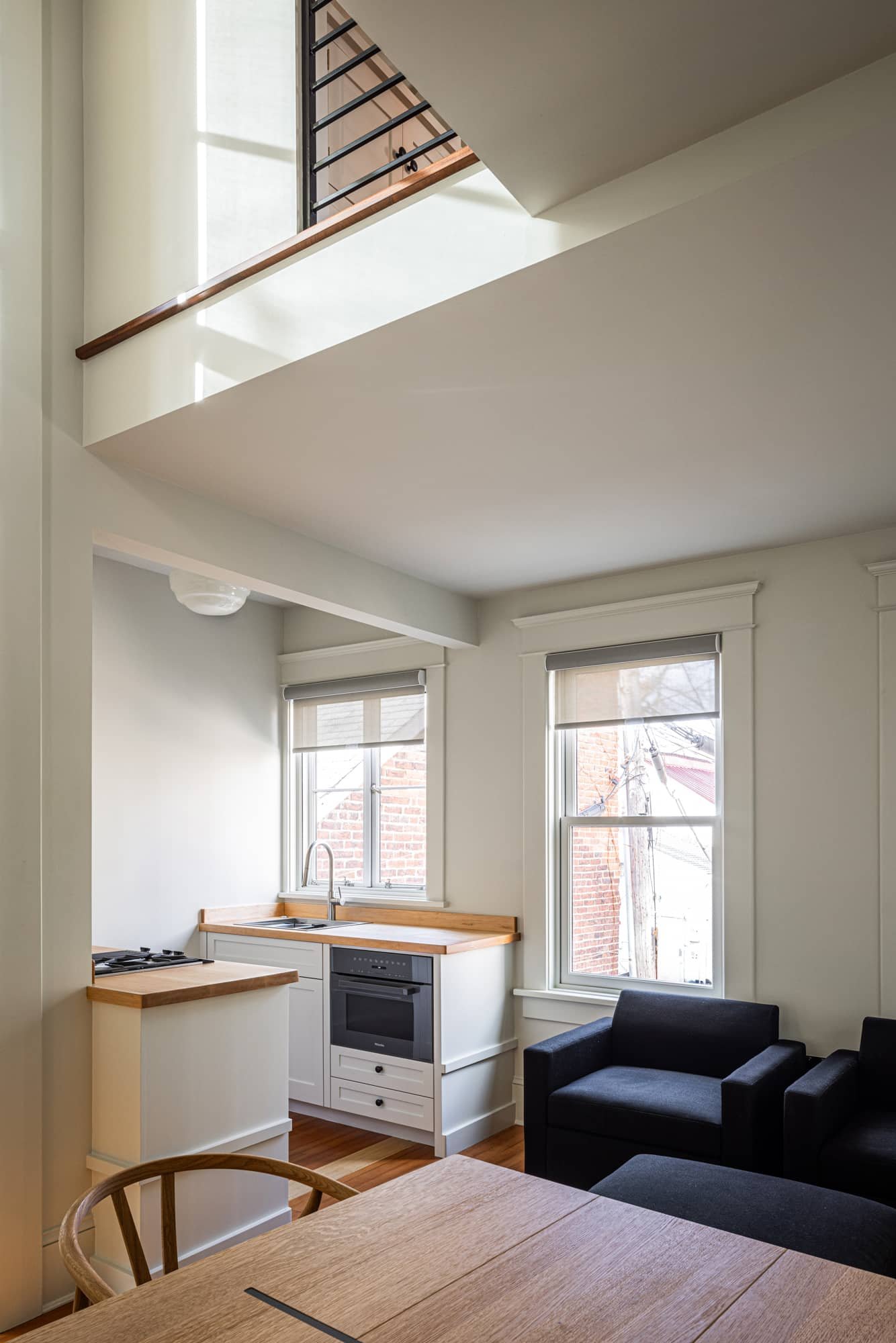 Interior view of a small kitchen and living room area with large windows, white walls, wooden countertops, a black sofa, and a wooden dining table.