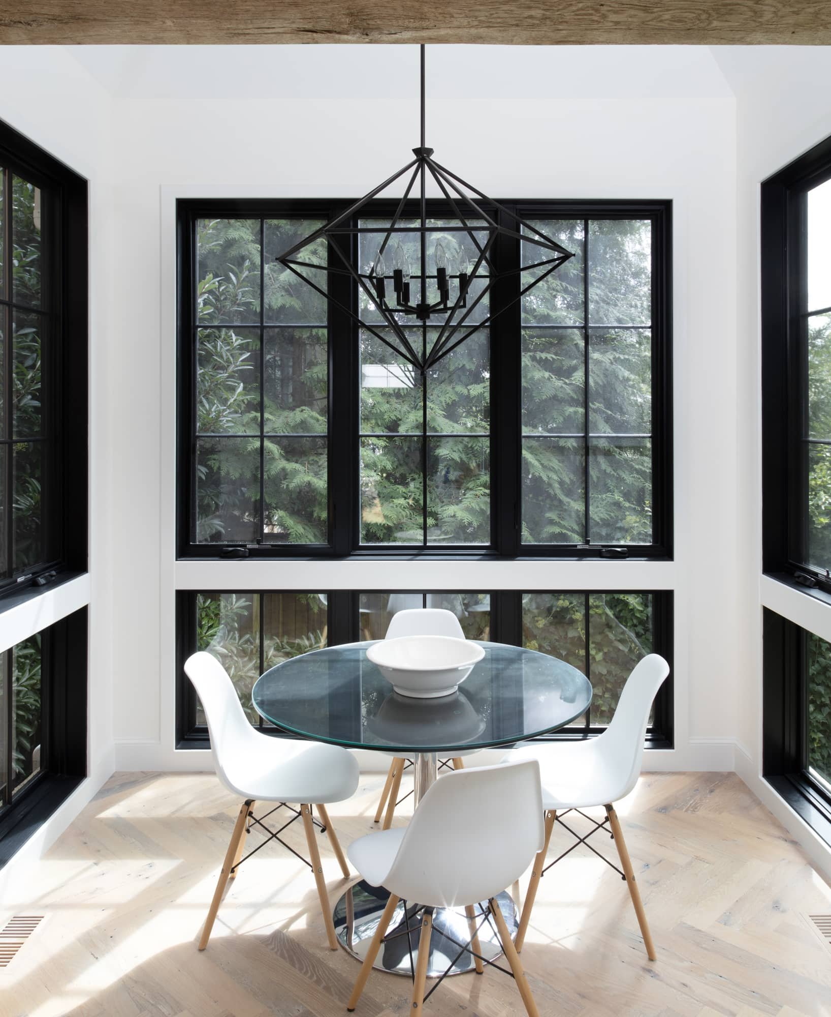 A modern dining area with a glass round table, four white chairs with wooden legs, a black geometric chandelier, and large black-framed windows showing greenery outside.