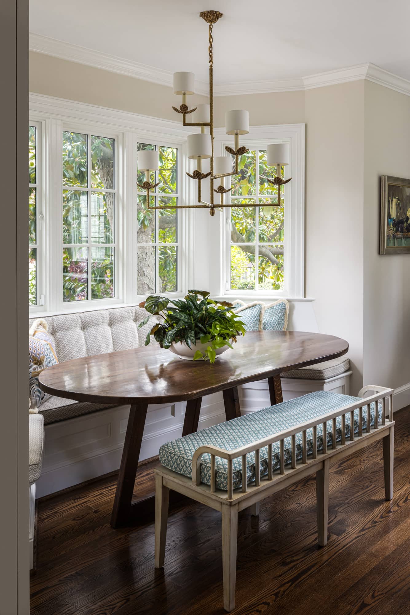 Dining nook with a wooden table, a white bench with patterned cushion, a potted plant centerpiece, large windows,. A modernized take on a classic style.