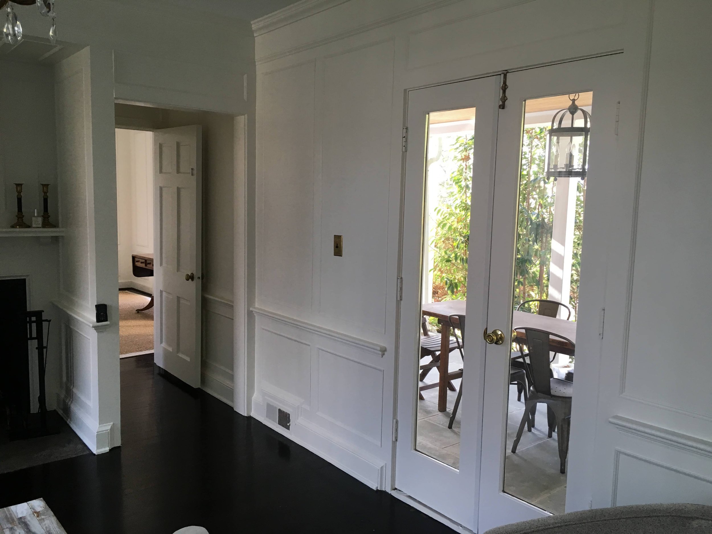 Interior view of a living room with white walls, hardwood floor, white double doors leading to an outdoor patio, and a chair partially visible at the bottom