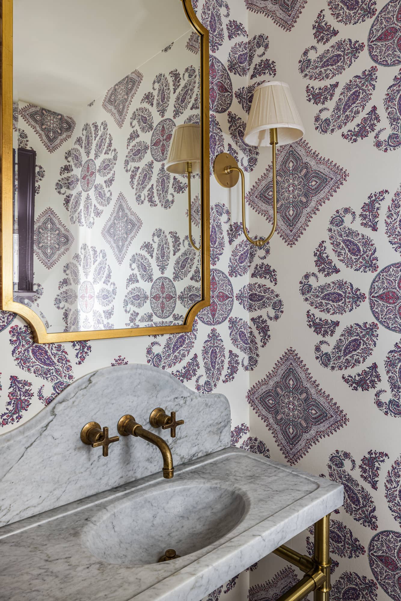 A bathroom with a marble sink, brass faucet and fixtures, a mirror with a gold frame, a wall-mounted lamp, and patterned wallpaper with purple and white design.