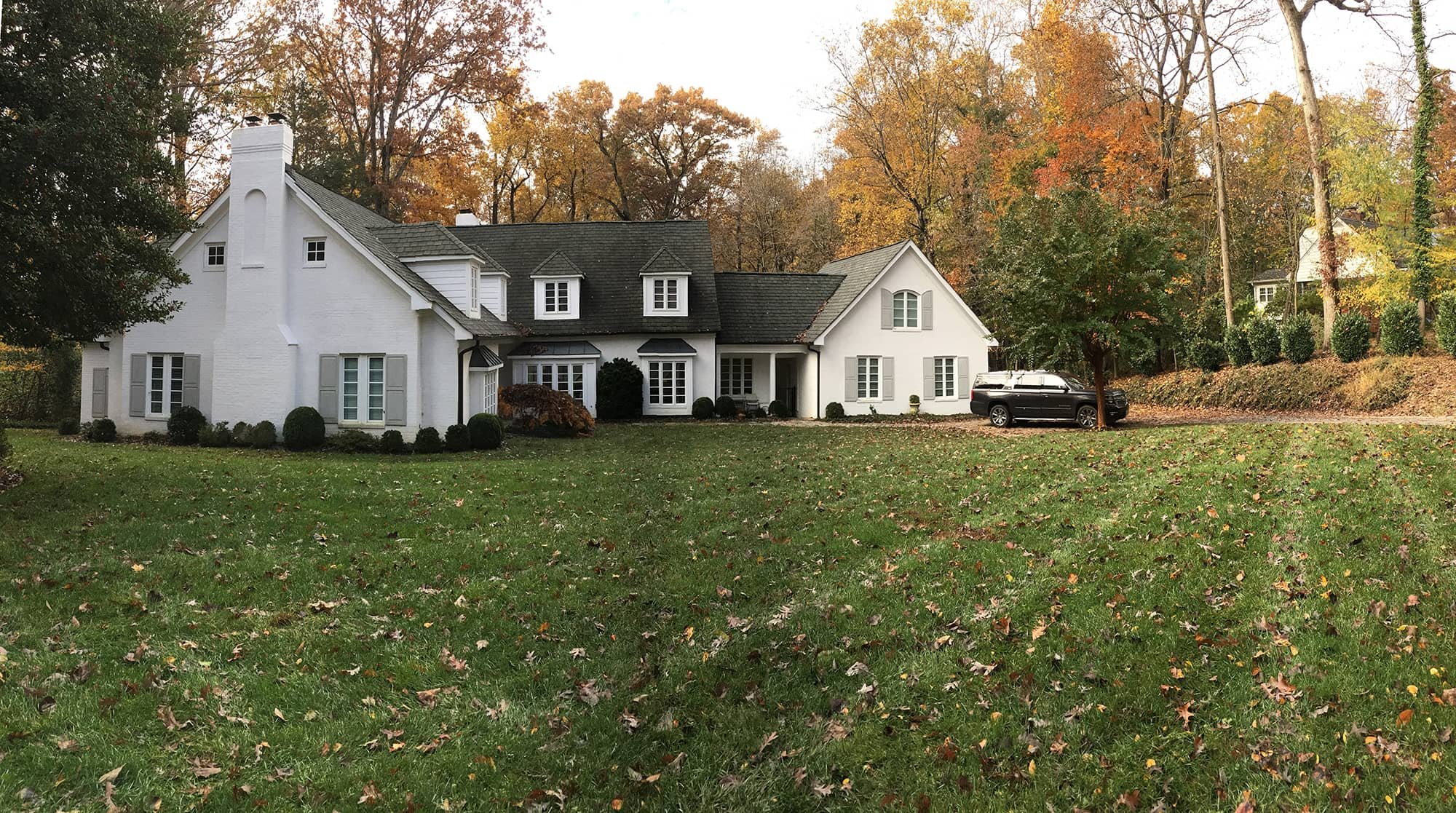 A large white house with multiple windows and a black roof, situated on a lawn with fallen leaves. There is a black SUV parked on the driveway on the right side. Autumn trees with yellow and orange foliage surround the house on a clear day.
