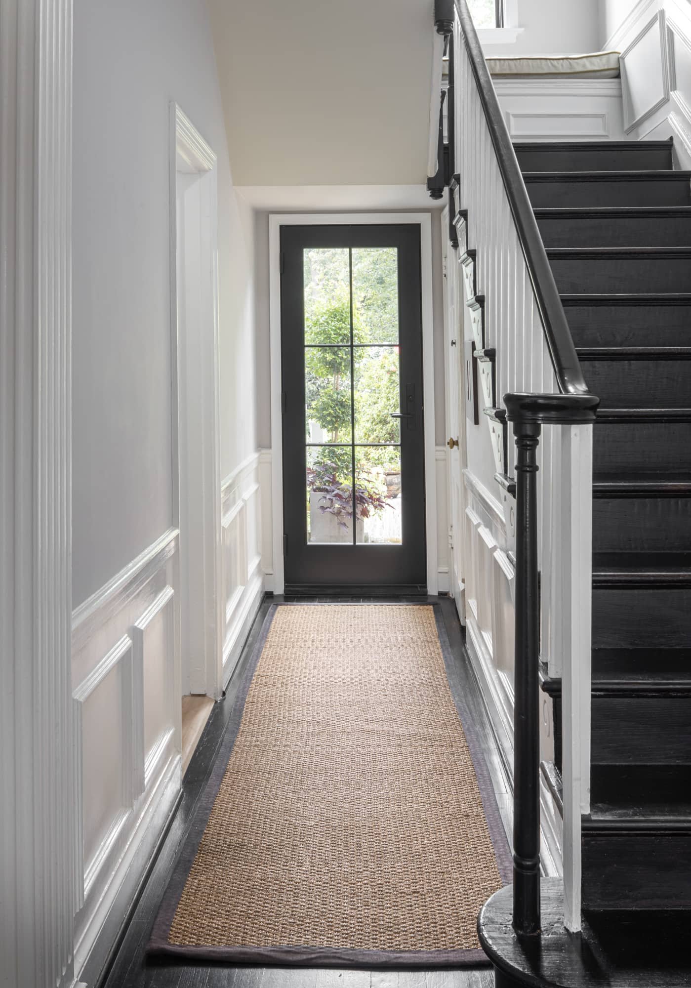 Interior of a house entryway with a black front door and a long beige rug on dark hardwood floor, with staircase on the right and white wainscoting on the walls.